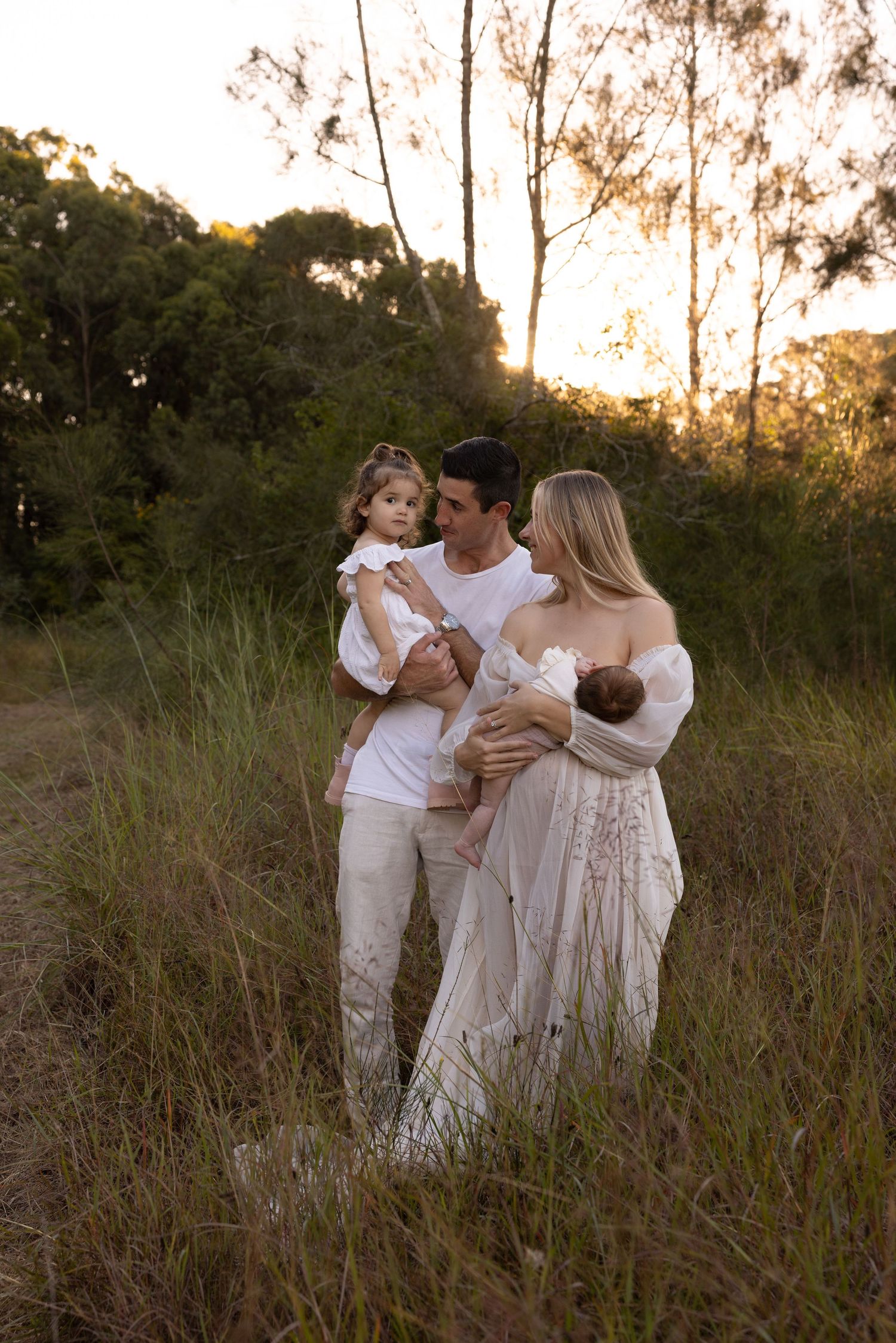 Family in white outfits embraces together in tall grass during golden sunset hour.