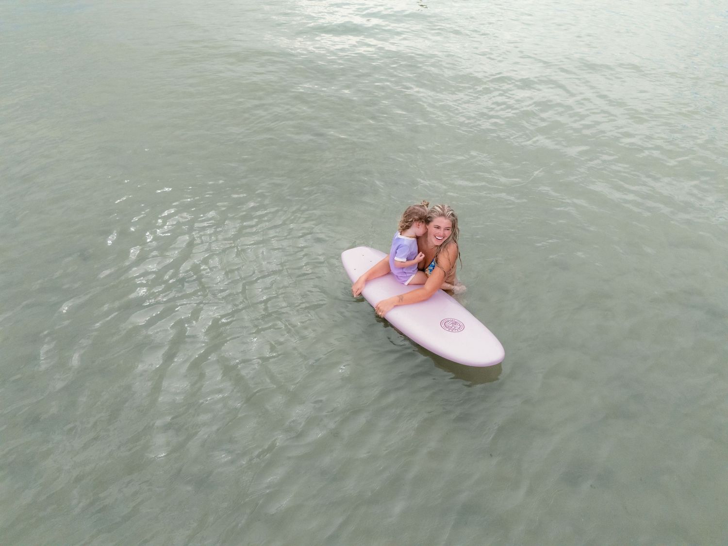 Aerial view of a surfer sitting on a pink and white surfboard in calm green waters.