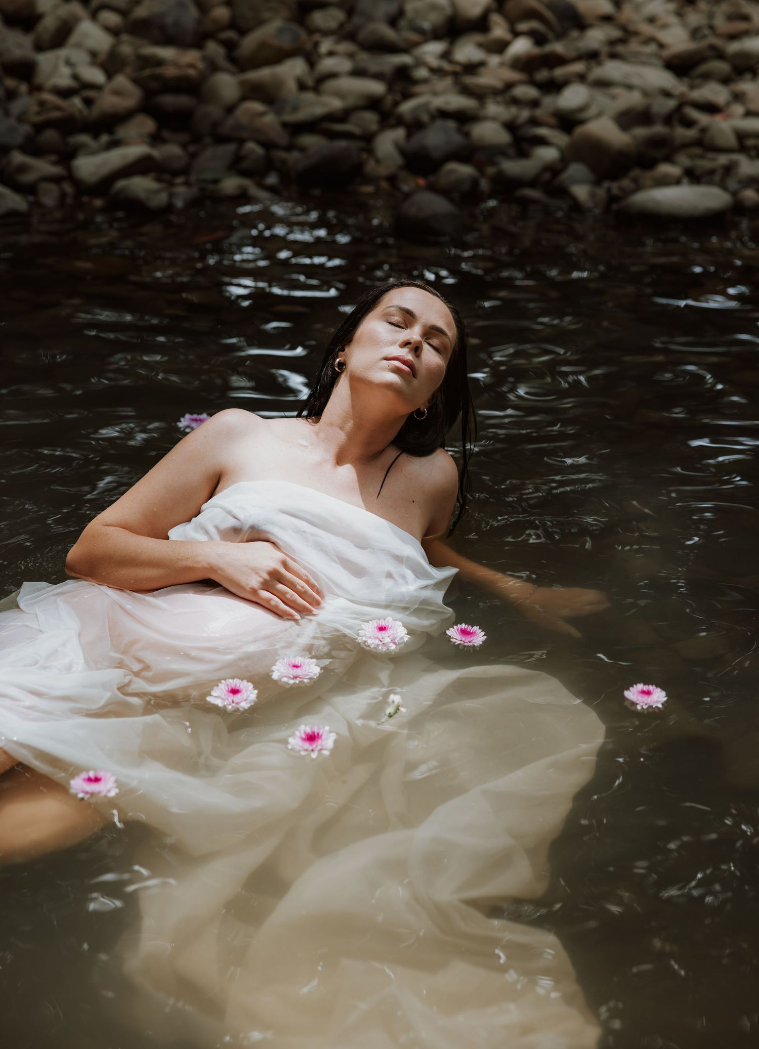 Maternity mother lays in the shallow creek water wrapped in fabric and near floating pink flowers.