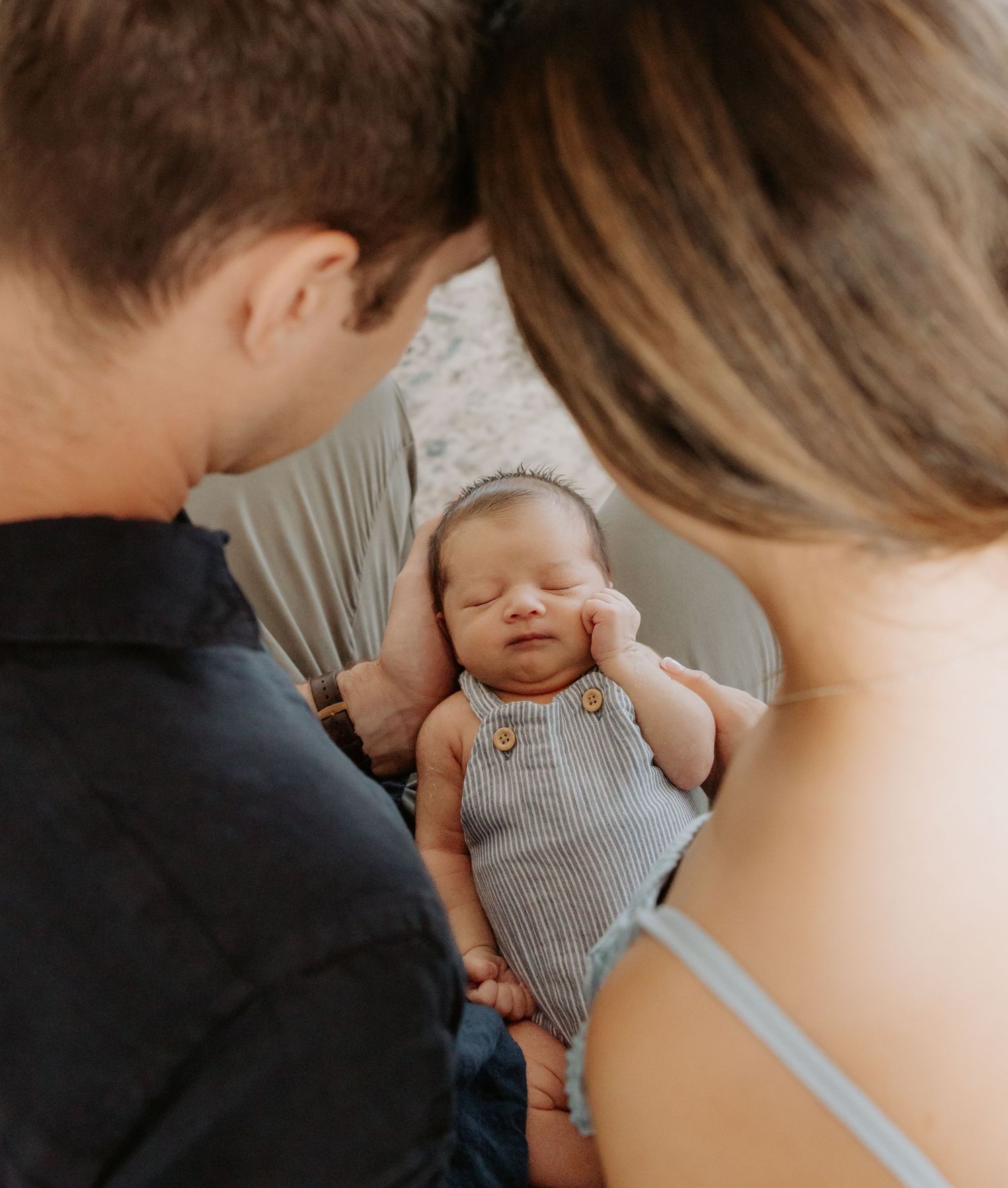 Mother and father lean towards each other, perfectly framing their newborn baby who is sleeping in their hands.