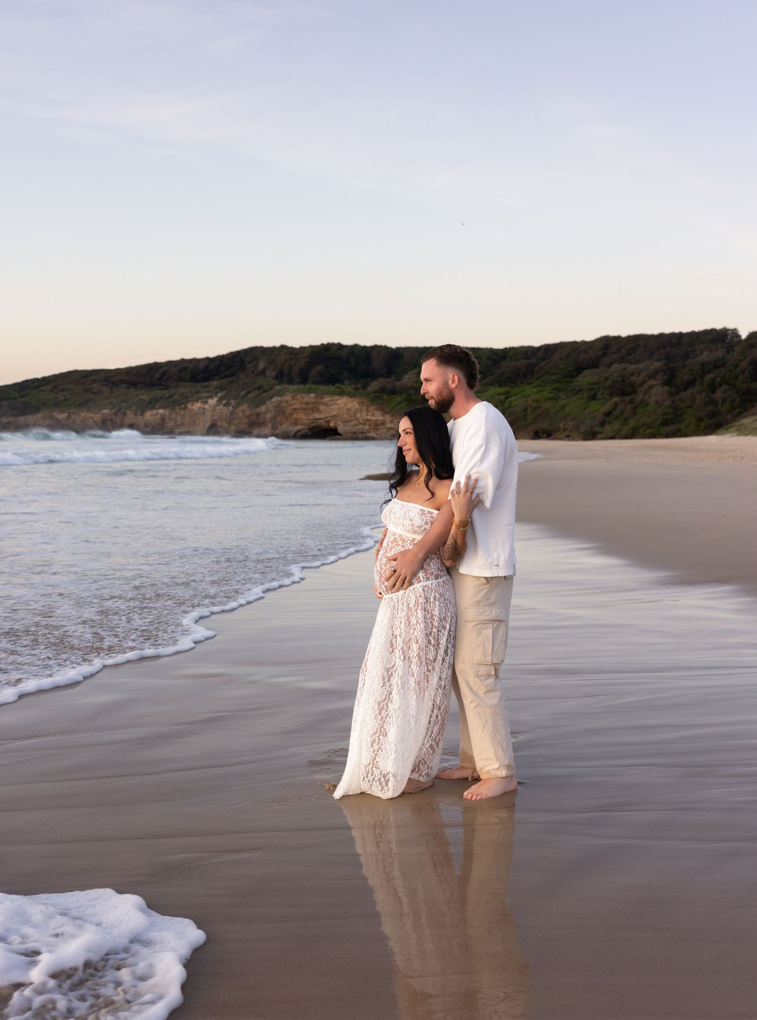 Expecting couple standing together at the shoreline during a maternity session on a Newcastle beach.