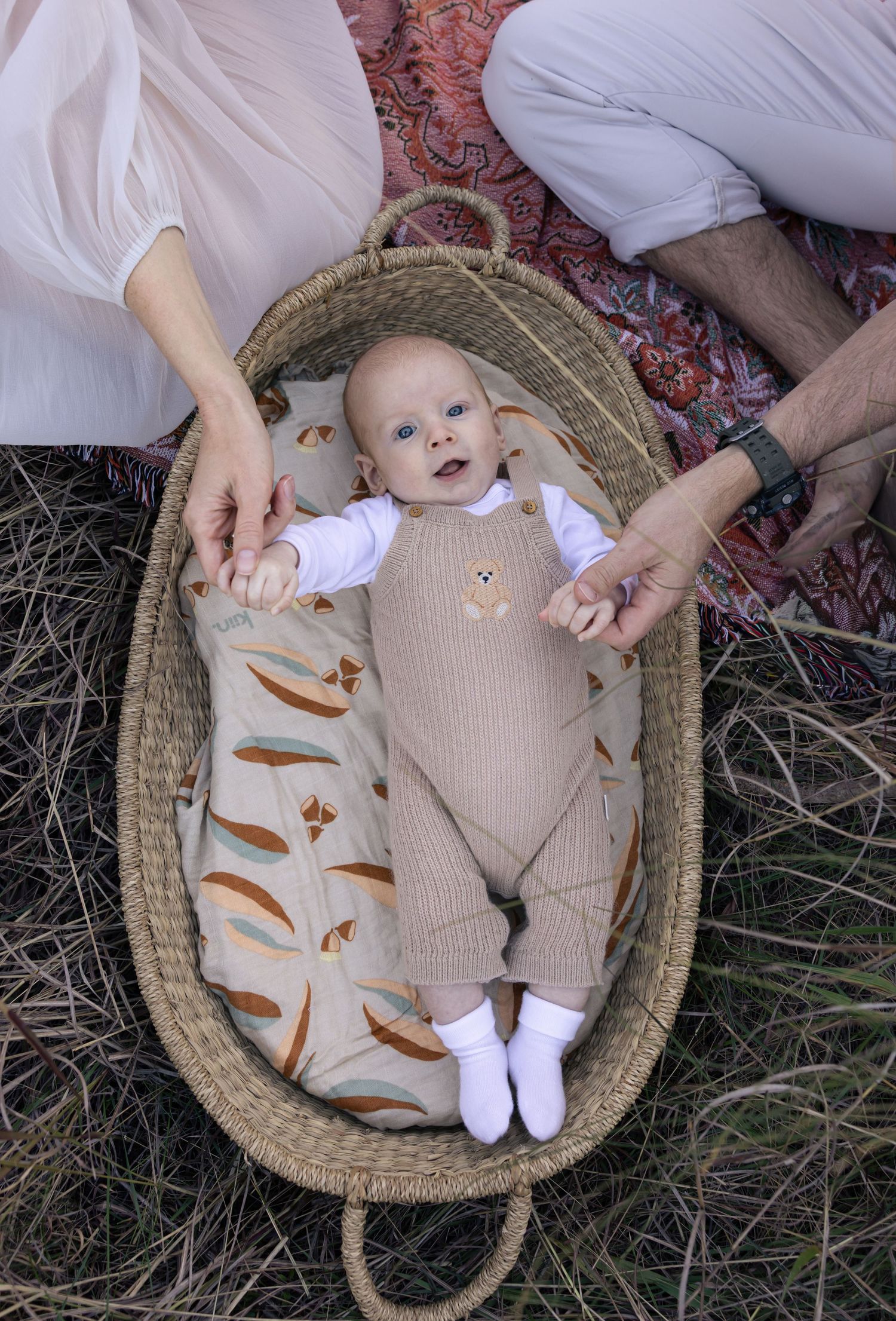 Young baby holds mum and dad's hands while laying in a baby basket in the grass fields at Booragul.