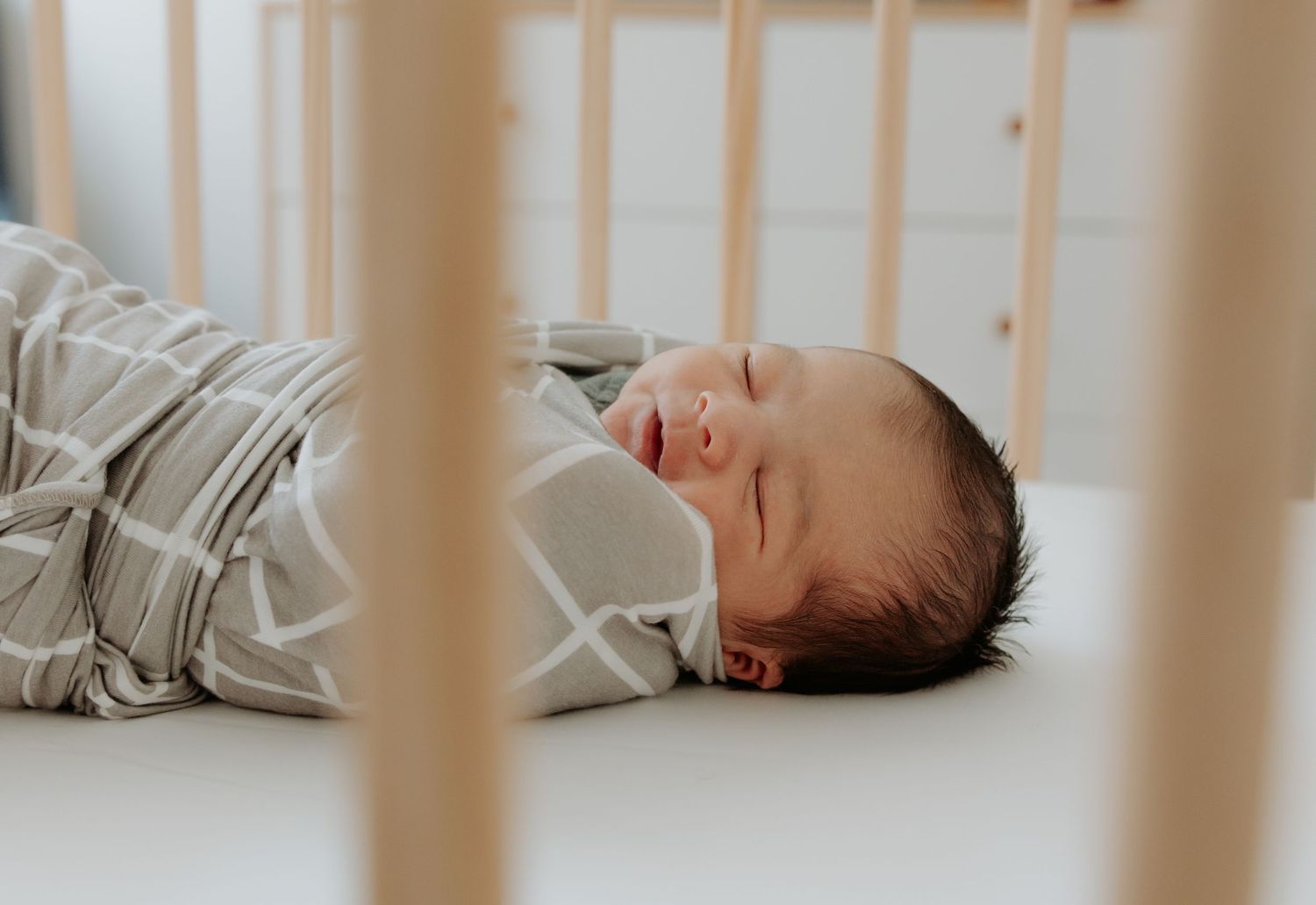 Newborn baby boy pictured through his timber cot, wrapped in a muslin wrap soundly asleep, white noise playing in the background.