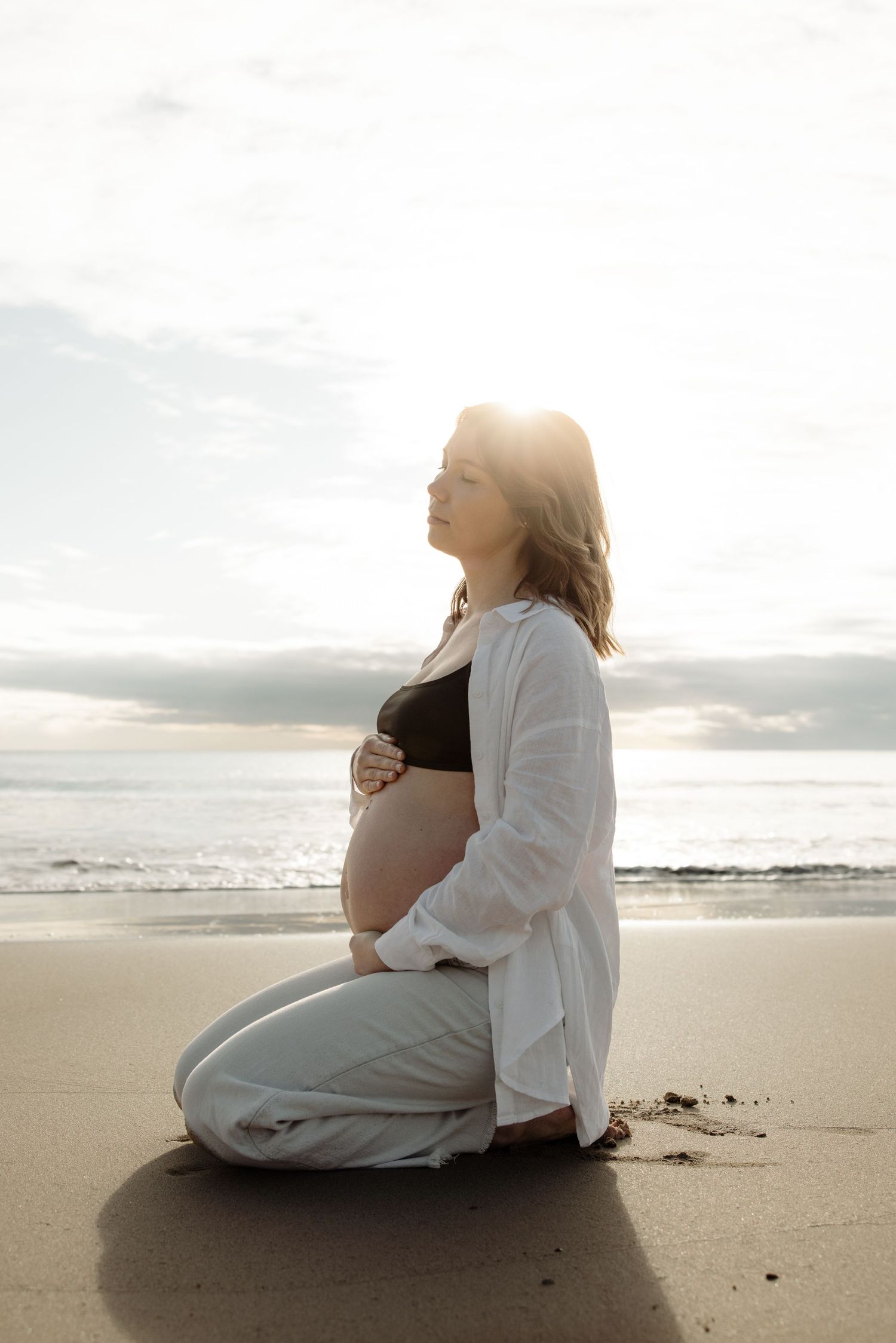 Pregnant mum sits in the sand and cradles her baby belly with clouds in the sky behind at a beach in Newcastle.