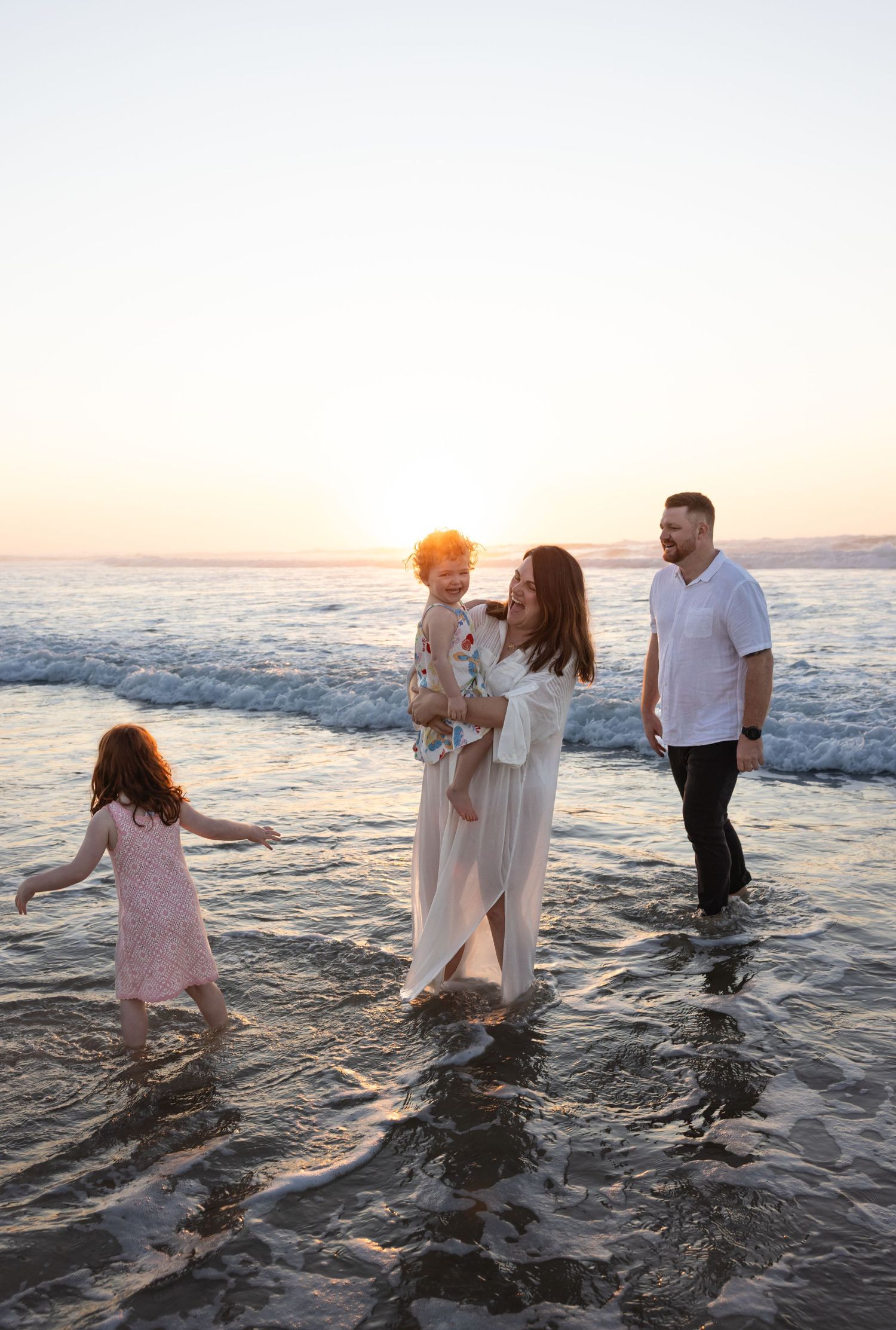 Family of four splash around in the shallow waters of Caves Beach with the sun rising over the ocean.
