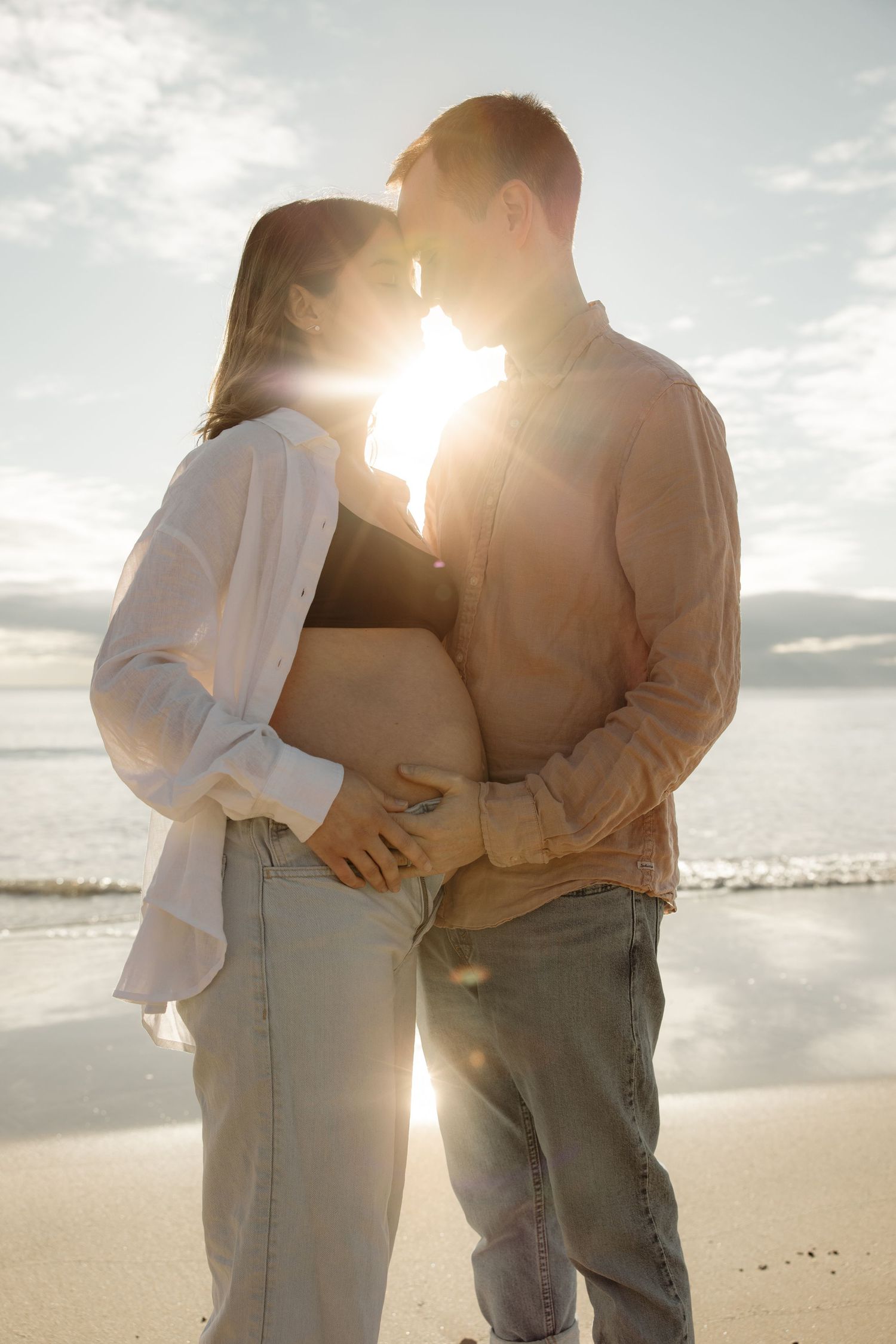 Expecting couple lean their heads together and close their eyes. The sun shines between them and reflects off the waters edge.