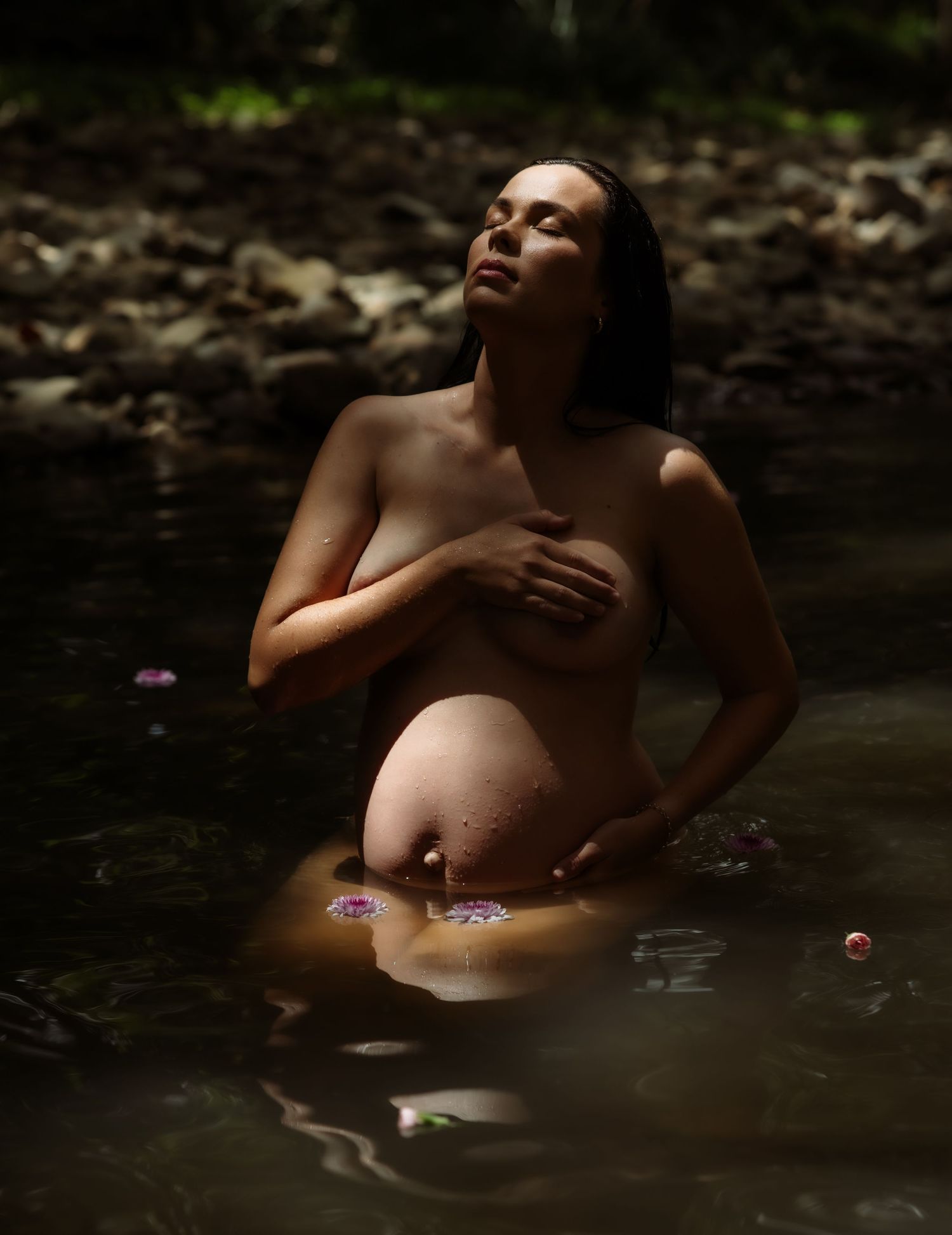 Pregnant mother sits in warm creek water with sunlight warming her face and belly.