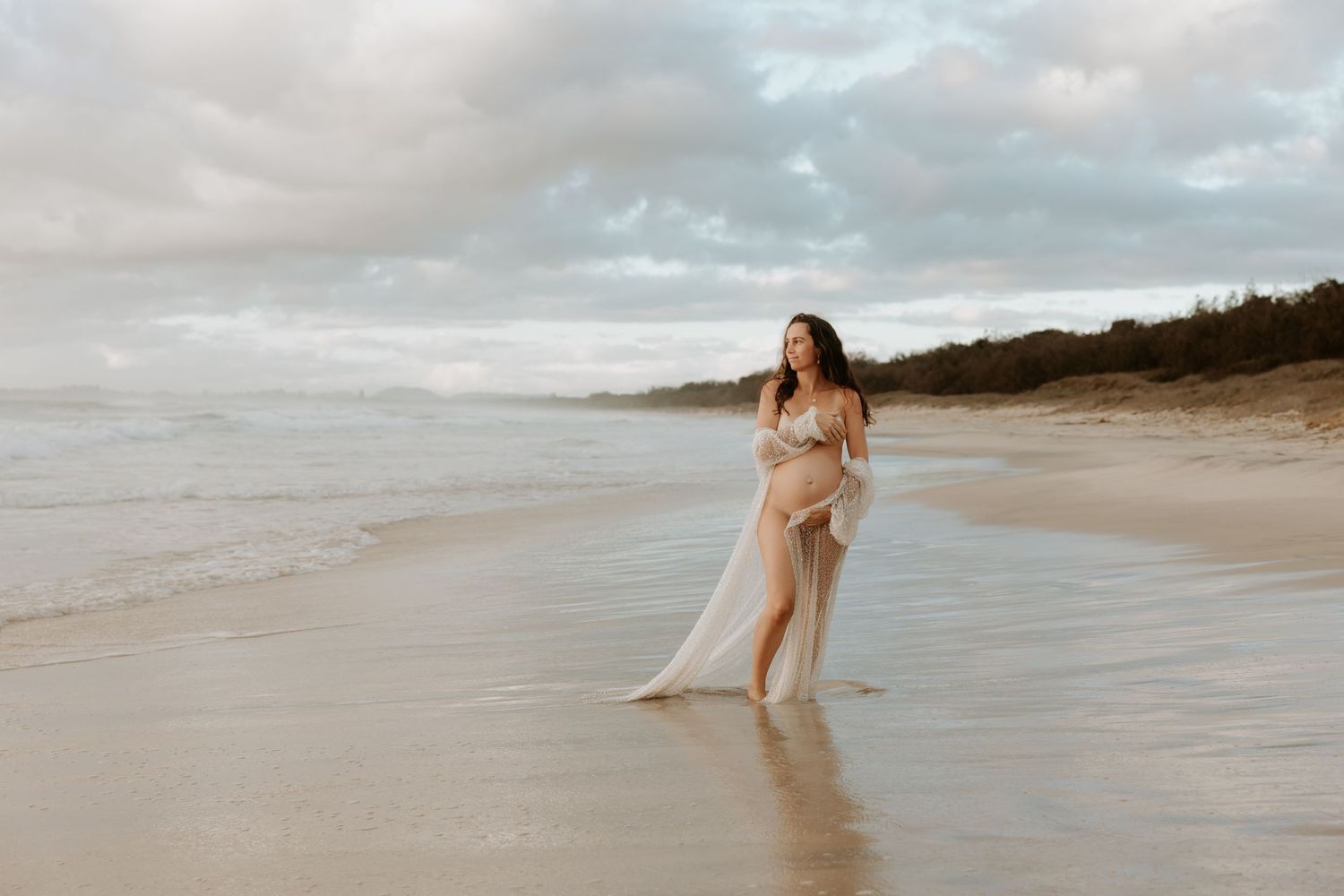 A girl in a flowy sequin dress poses gracefully along a serene beach shoreline at sunset.