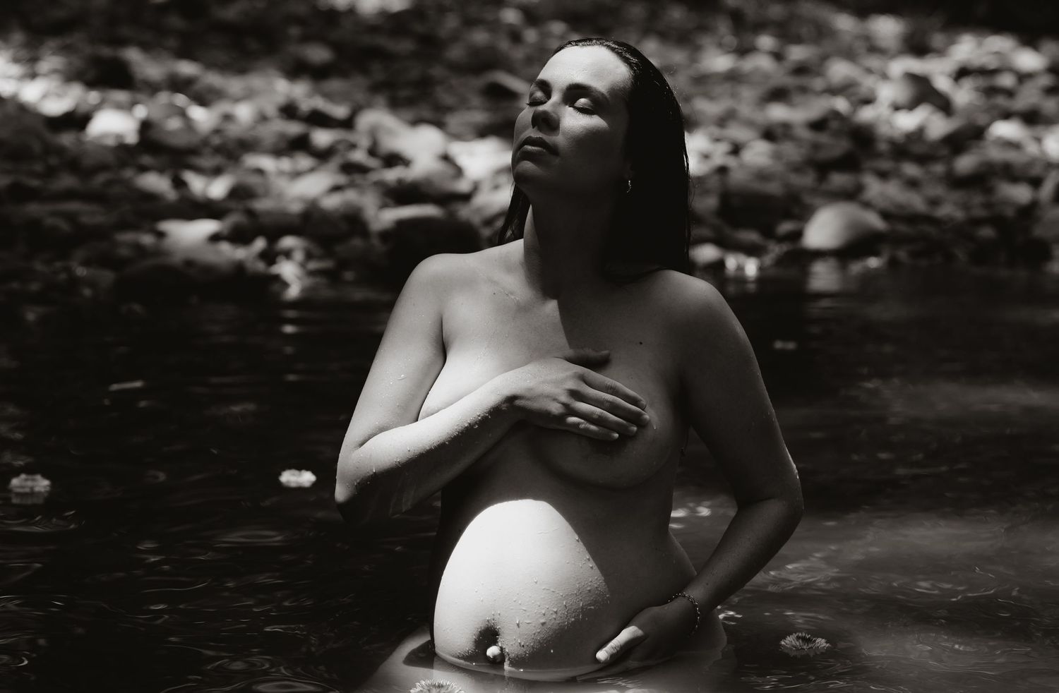 Pregnant mother sits in shallow water, looking up towards the dappled sunshine streaming in through the trees.