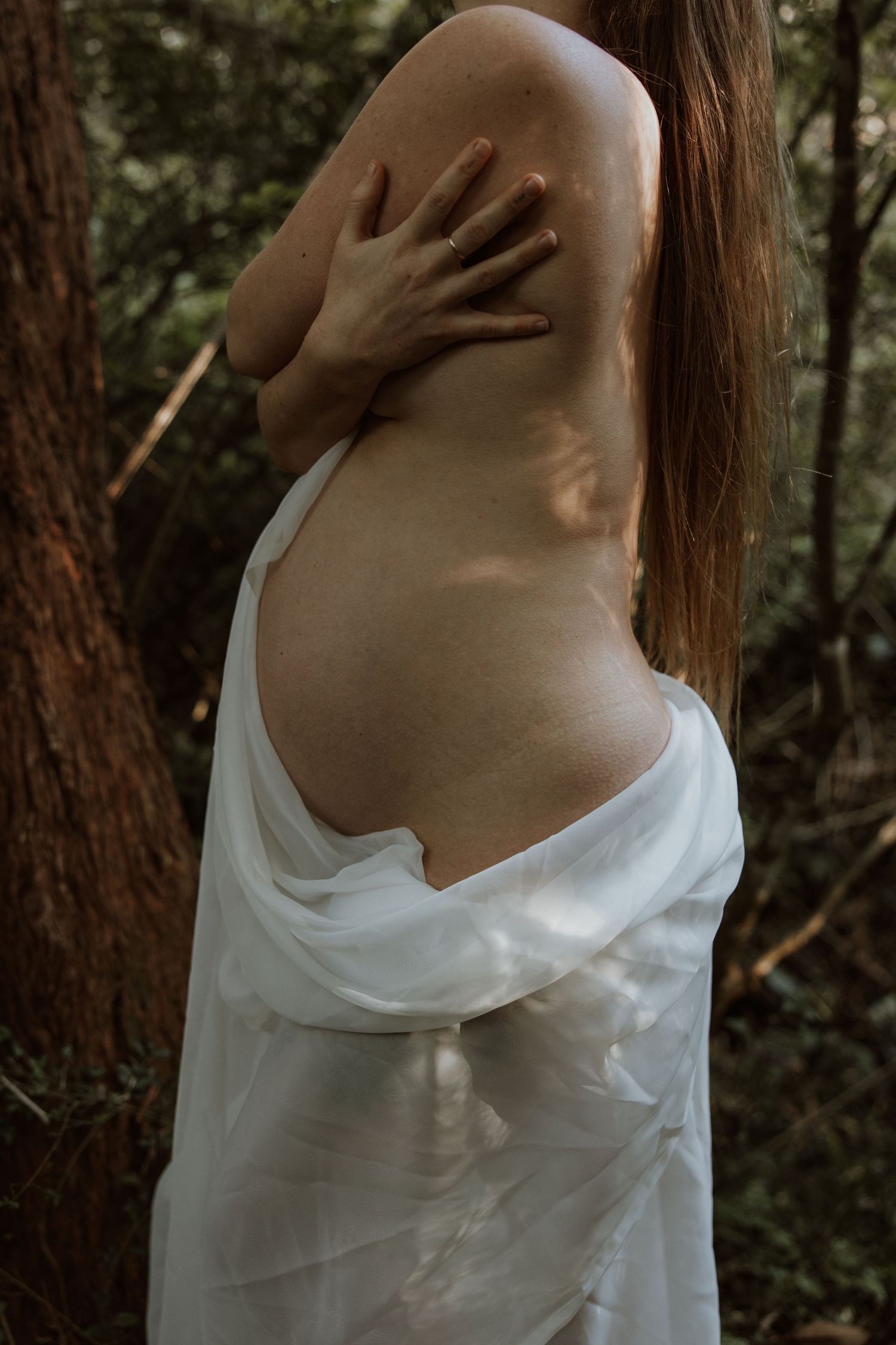 Maternity photoshoot in the bush at Glenrock with dappled light filtering through the trees. Mum holds herself in an embrace.