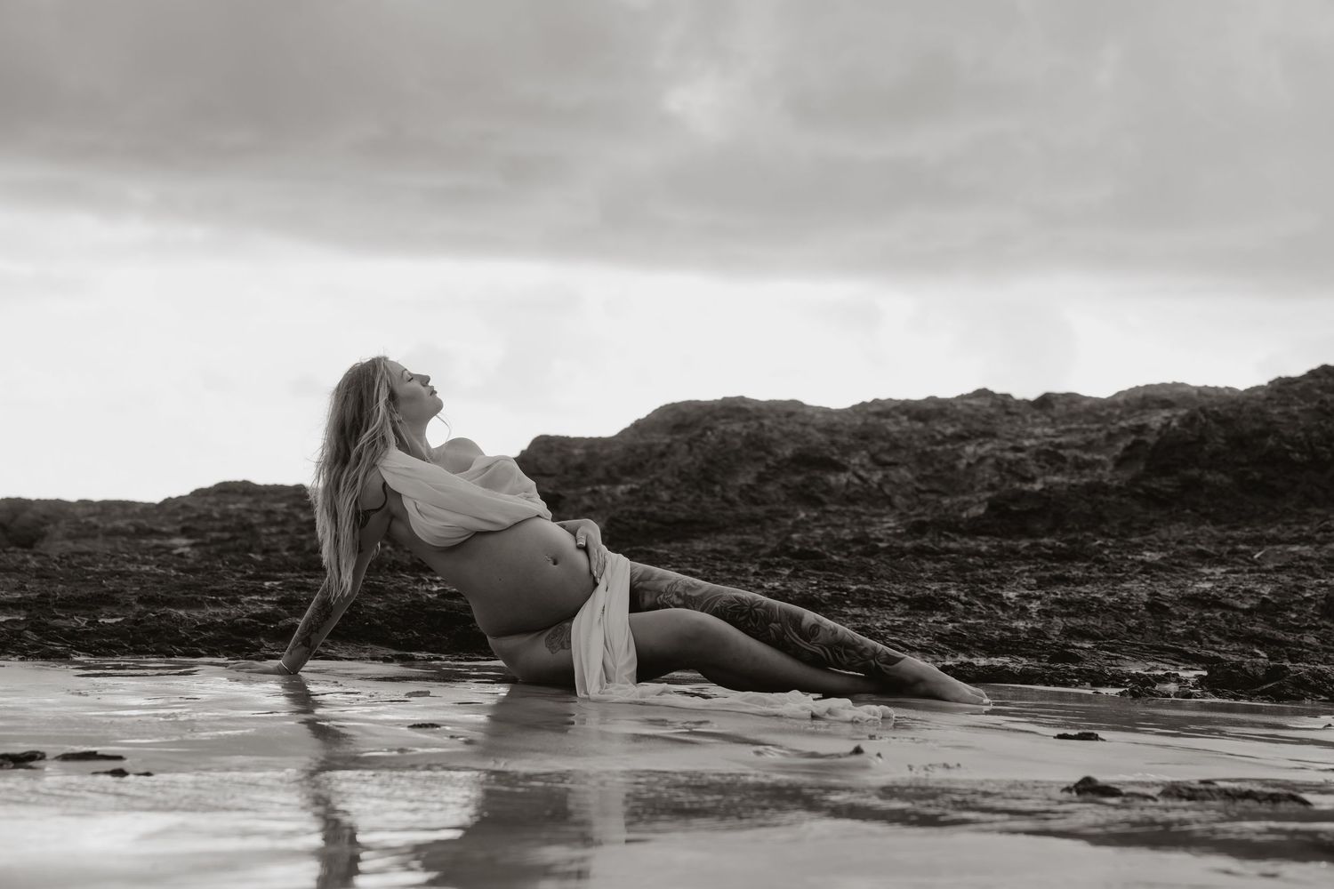 Tattooed pregnant mother holds growing belly as she reclines in shallow waters at the beach.