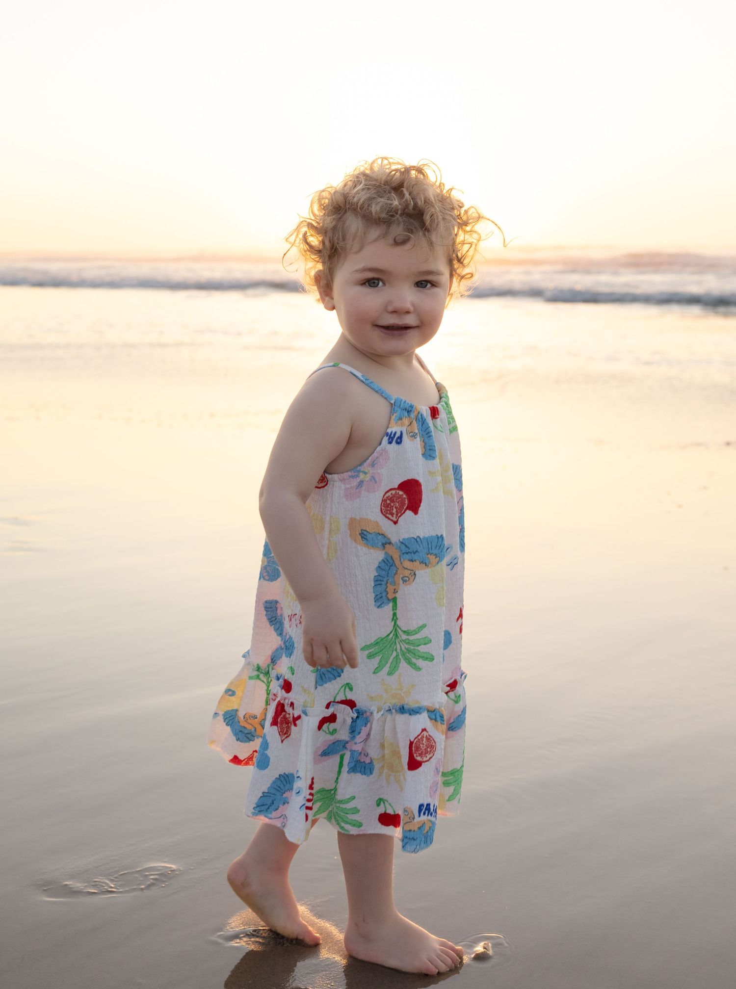 2 year old pauses at the beach, her curly hair glowing golden in the morning sunshine.