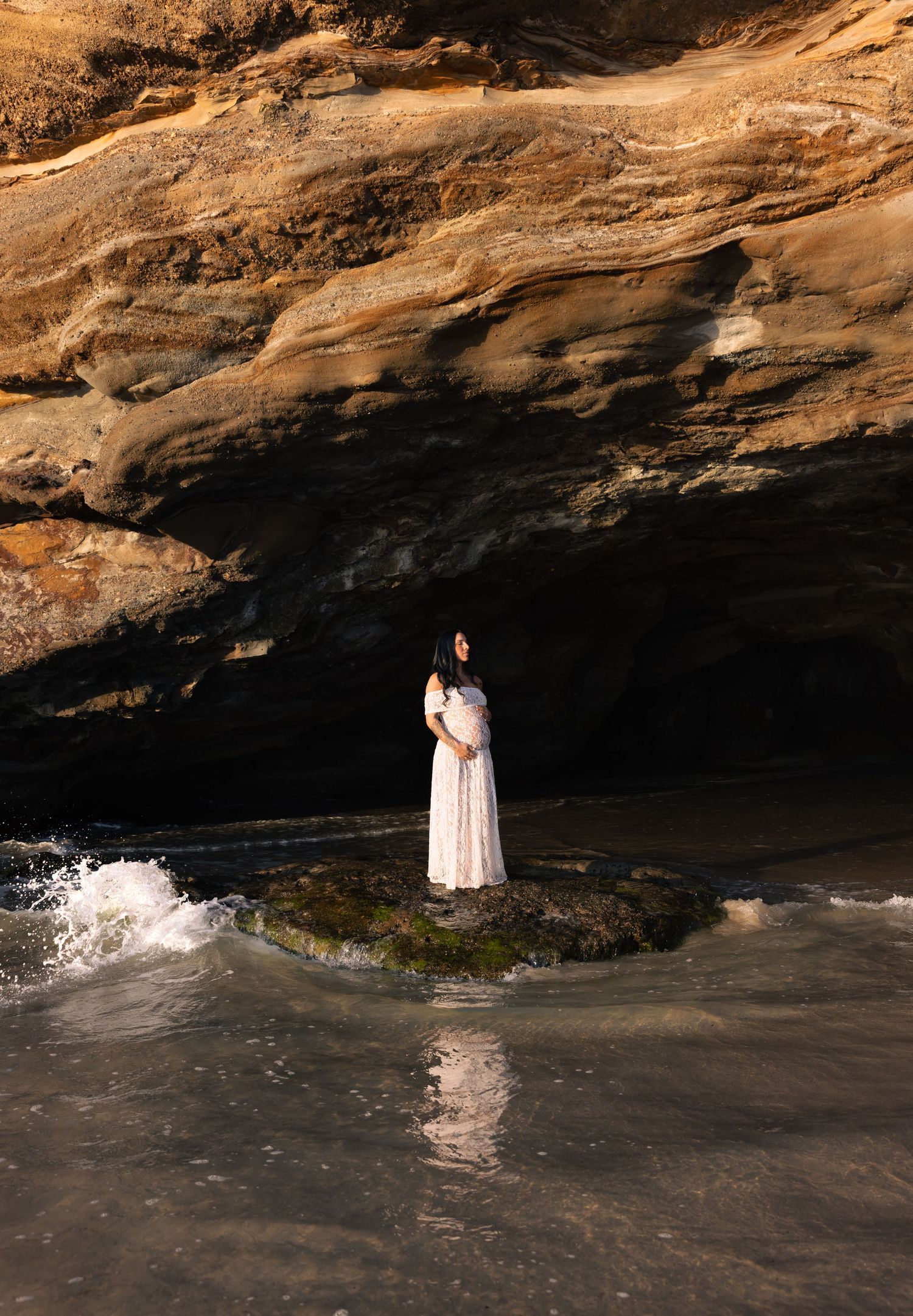 Woman in white dress stands on a rock as small waves gently roll below the rock.