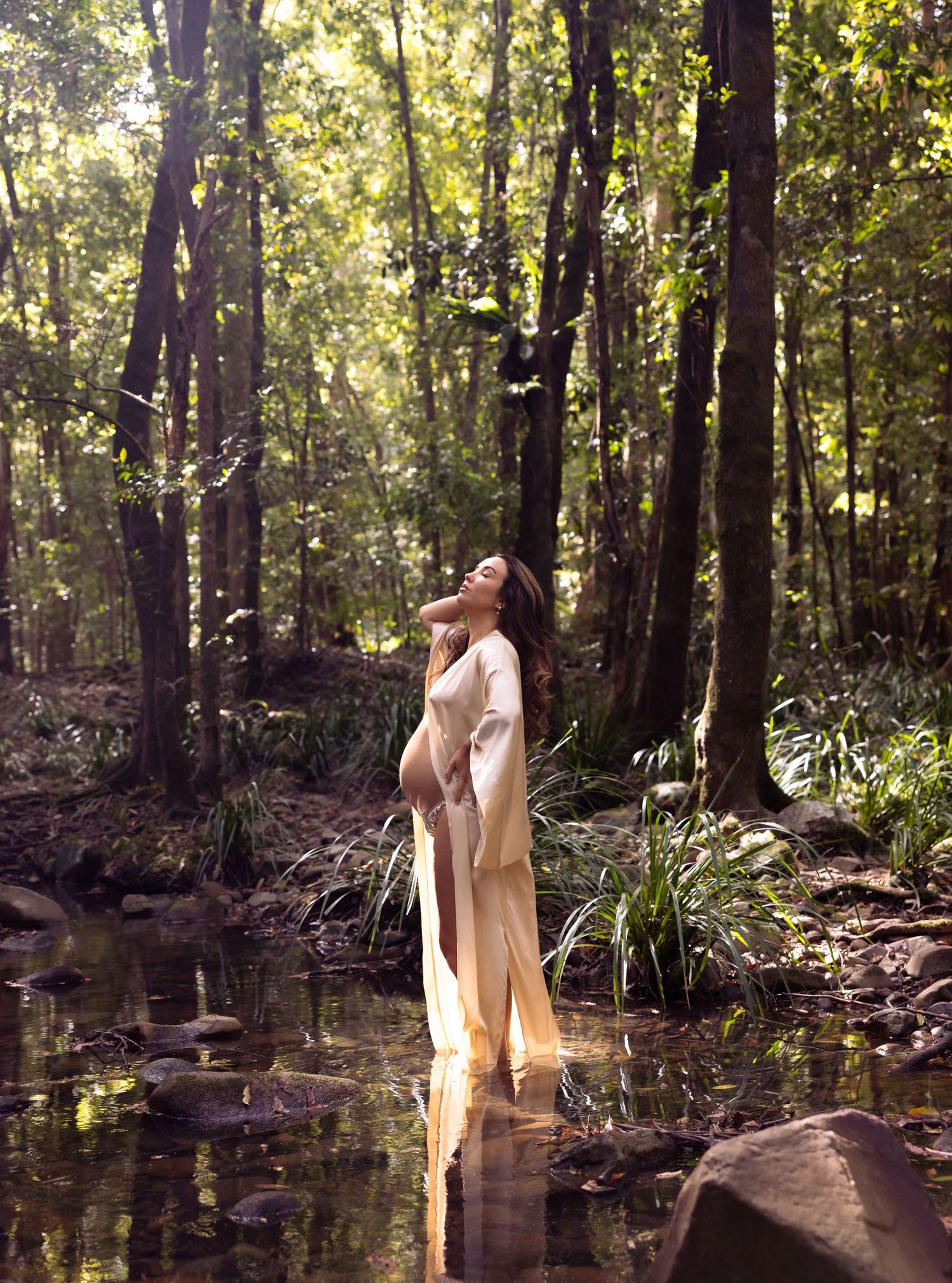 Pregnant mother stands in the cool creek as sunlight streams through leaves in the trees and lands on her face, illuminating her.