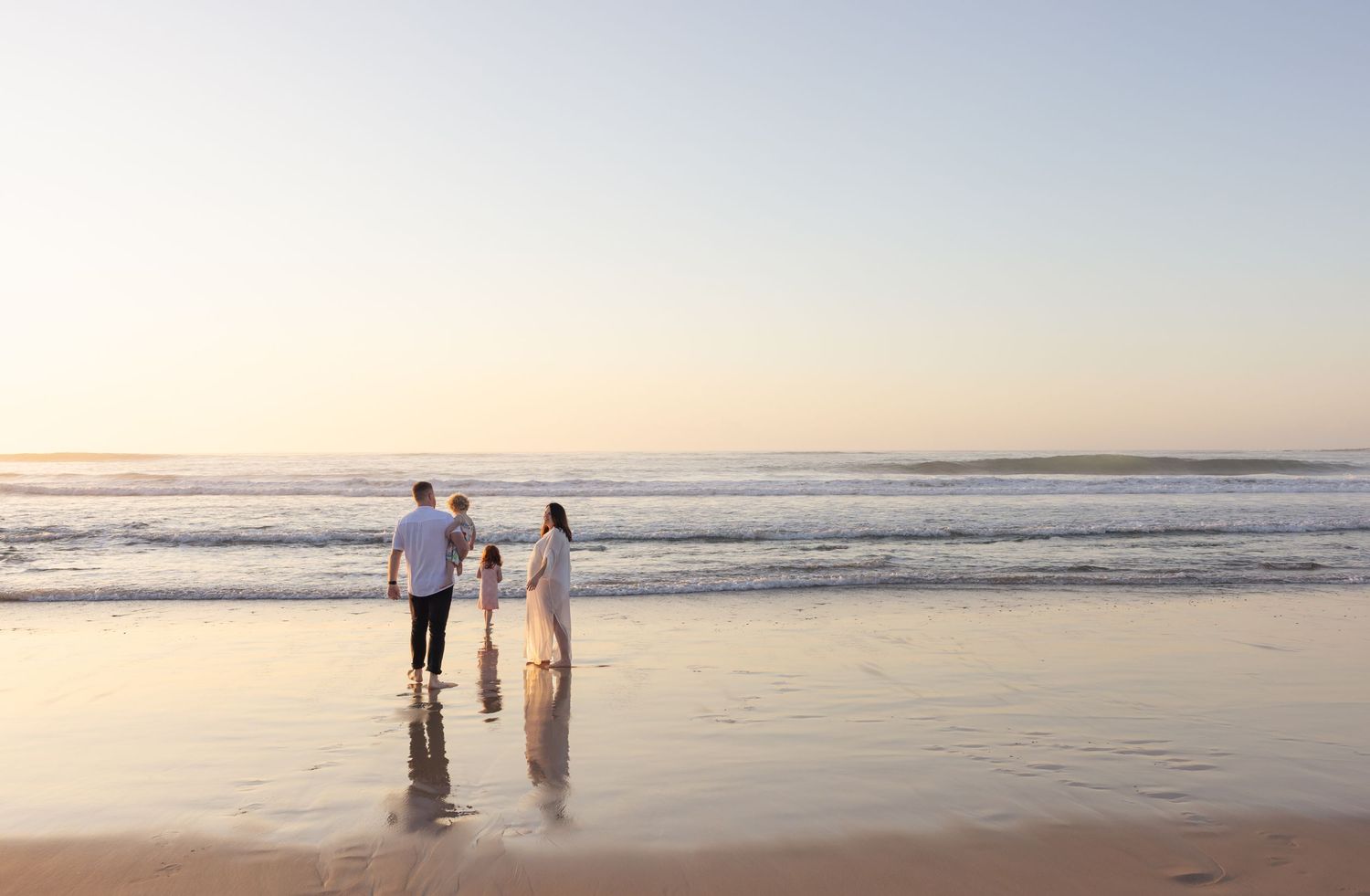 Family of four walking along the shallow water as ocean waves roll in and the sunrise casts reflections of the family in the water.