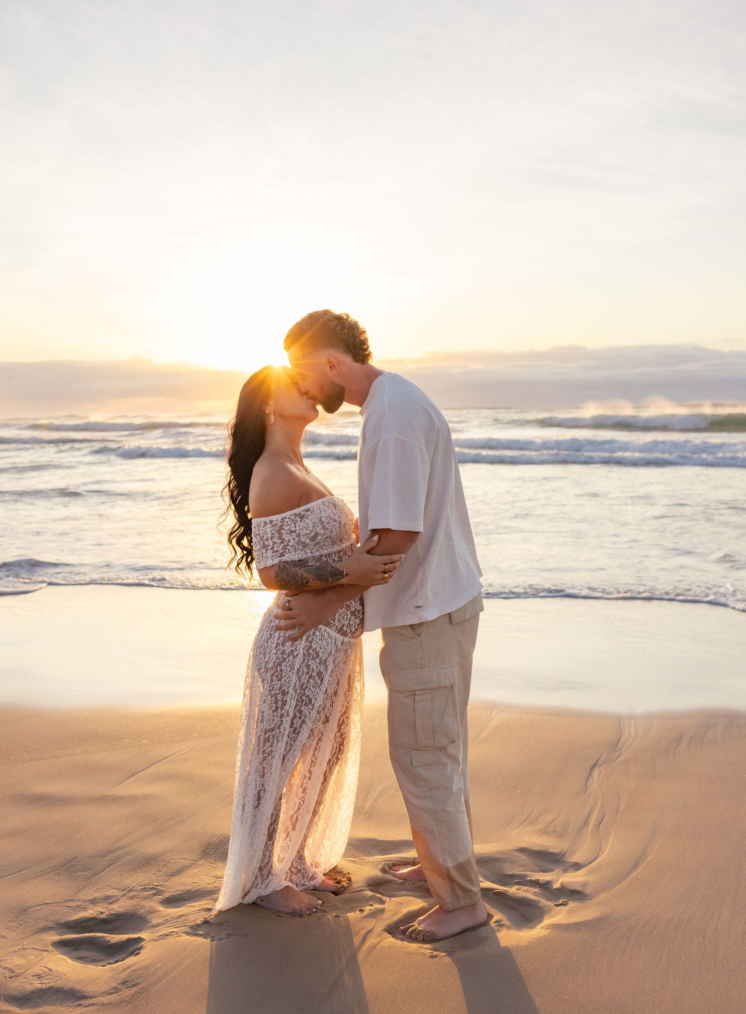 Maternity couple at the beach with the waves in the background and sunshine streaming in behind their heads creating a glow.
