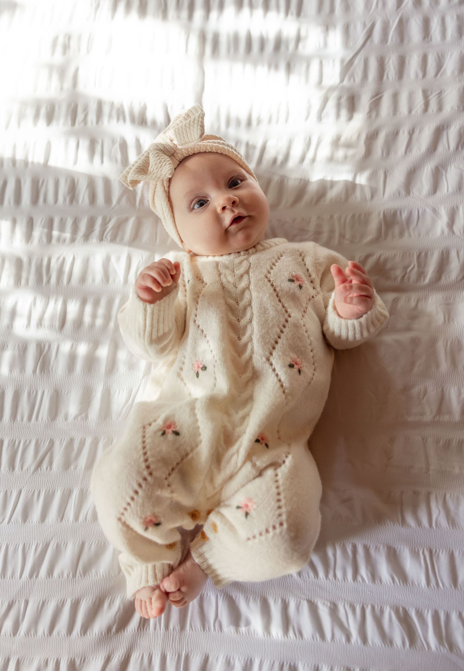 A baby wearing a cream-colored knit onesie with delicate embroidery lies on white bedding with sunlight streaming over the bed.