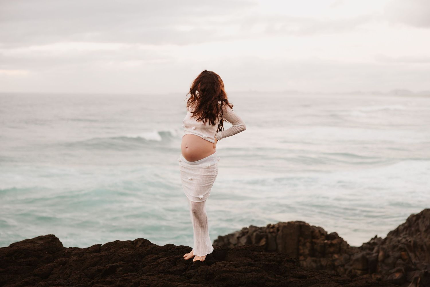 Pregannt mum stands on rocks looking over the ocean at Redhead beach, the wind in her hair.