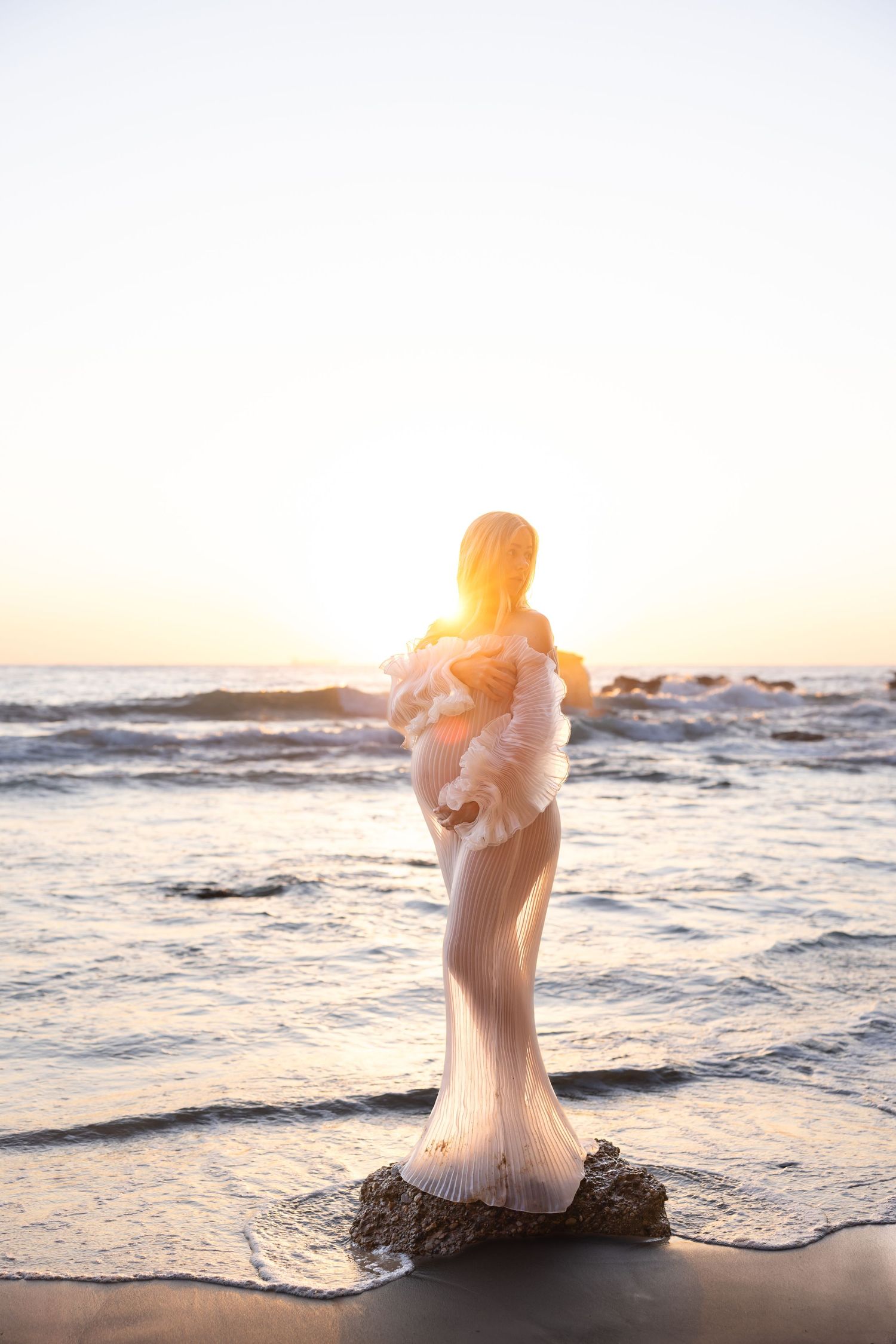 Pregnant mum stands on a rock with the sun rising behind her in a client wardrobe dress.