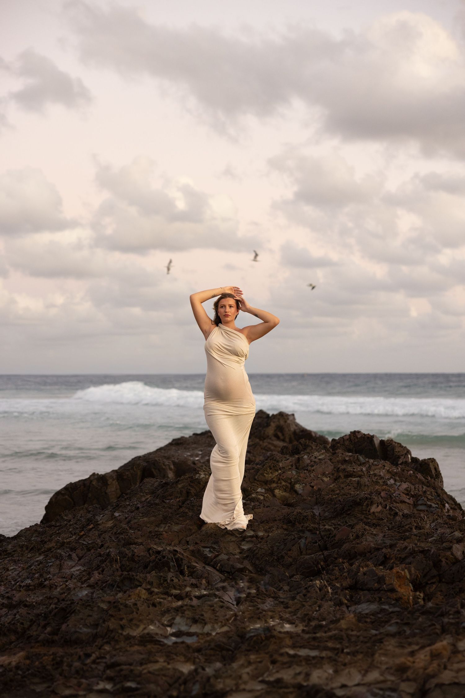 Pregnant mum stands on rocks with the ocean behind her as three birds fly in perfect formation behind her, framing her arms.