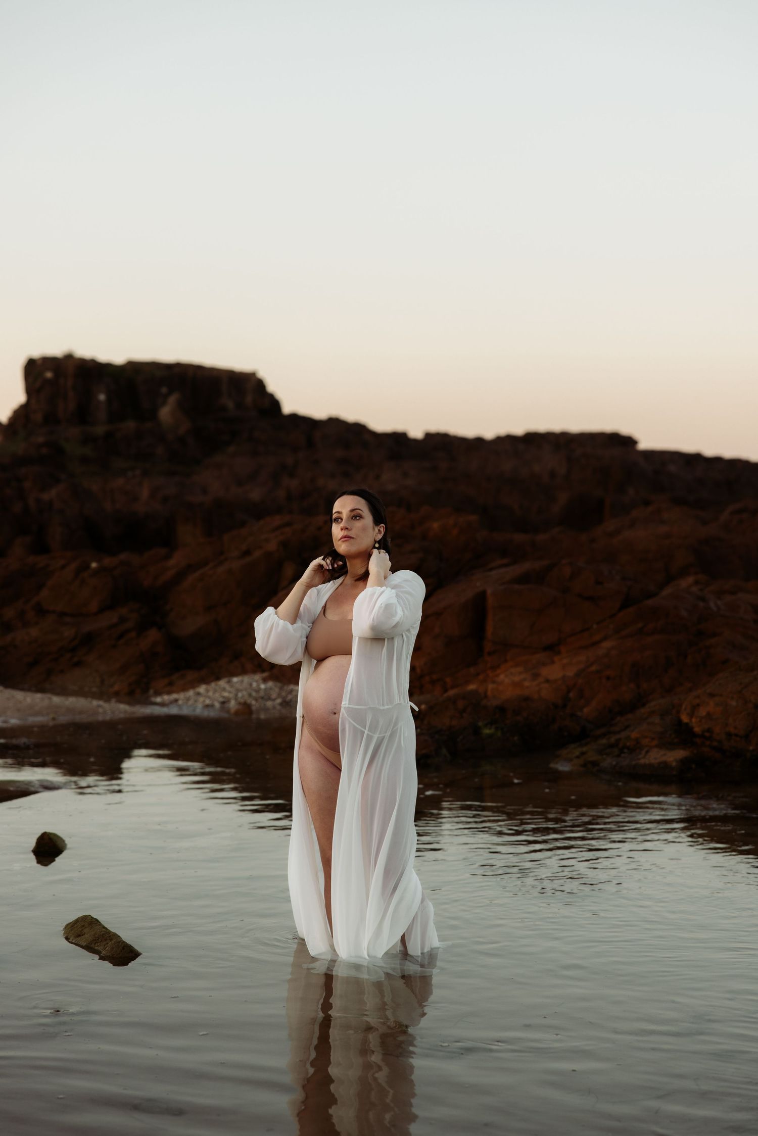 Pregnant mum stands in shallow waters, her hands smoothing her hair back.