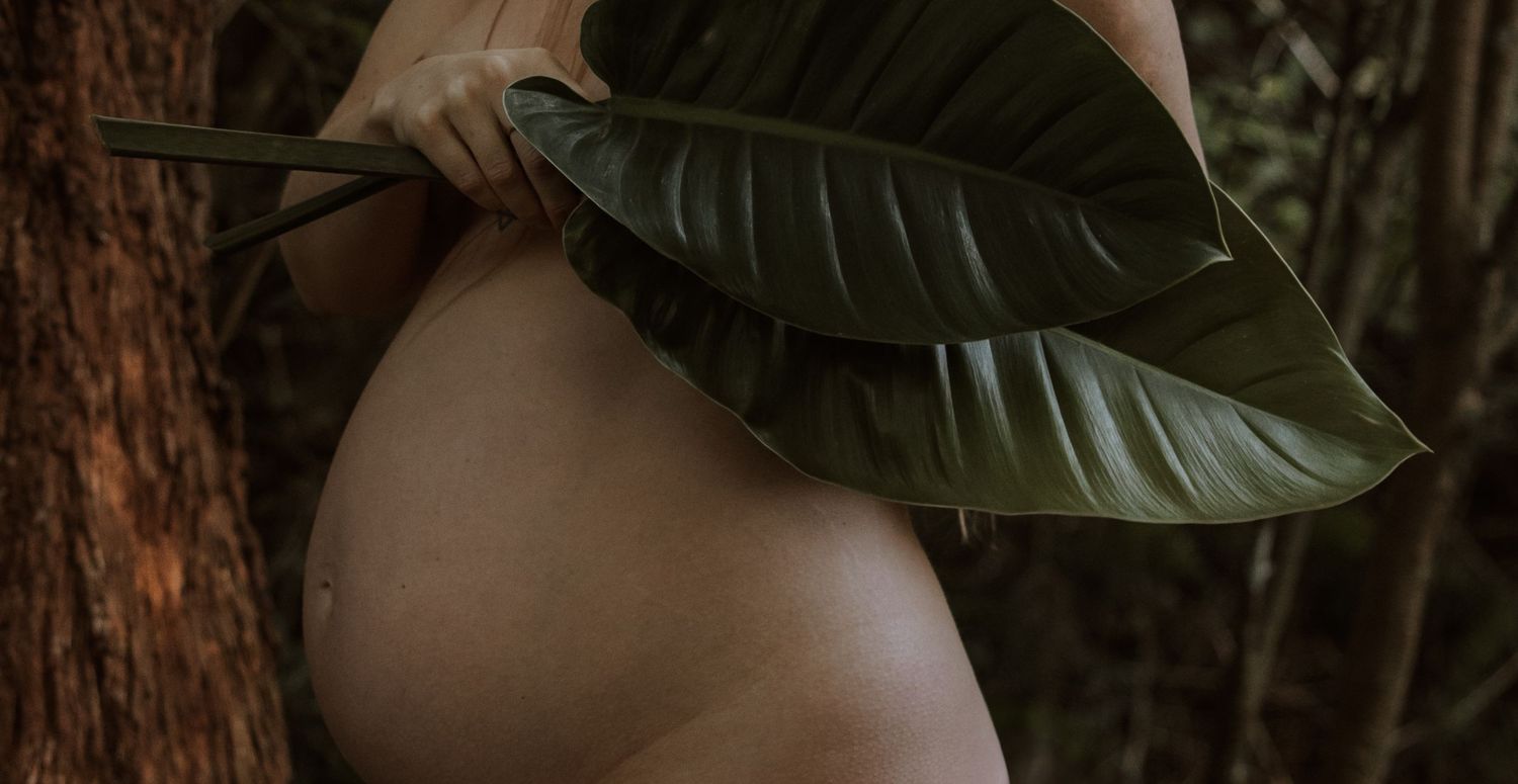 Mother holds large plant leaves over her pregnant belly with trees in the background in Newcastle.