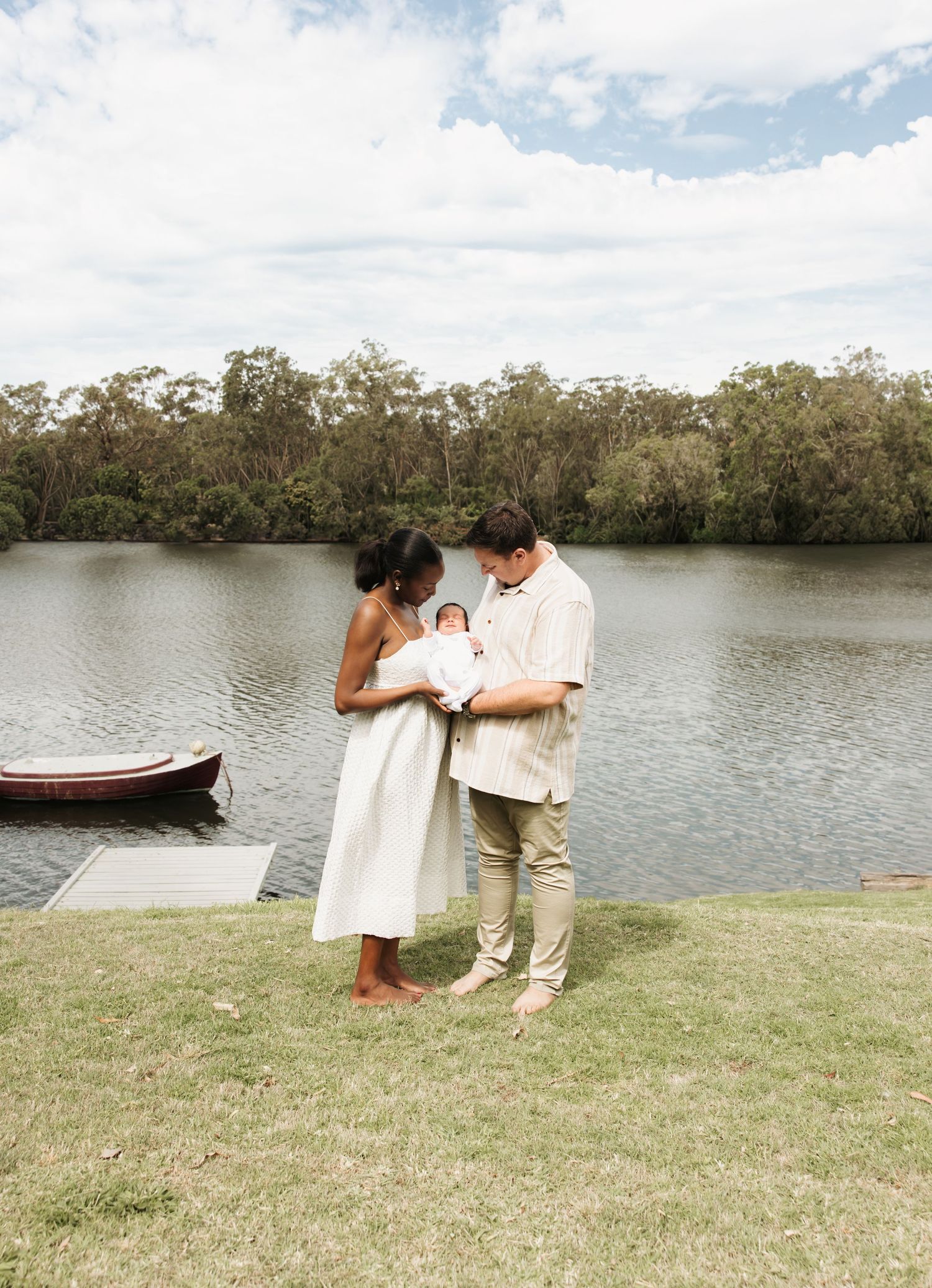 Mum and dad hold baby boy outside on backyard lawn with the creek behind them and a boat gently floating at its mooring in Lake Macquarie.