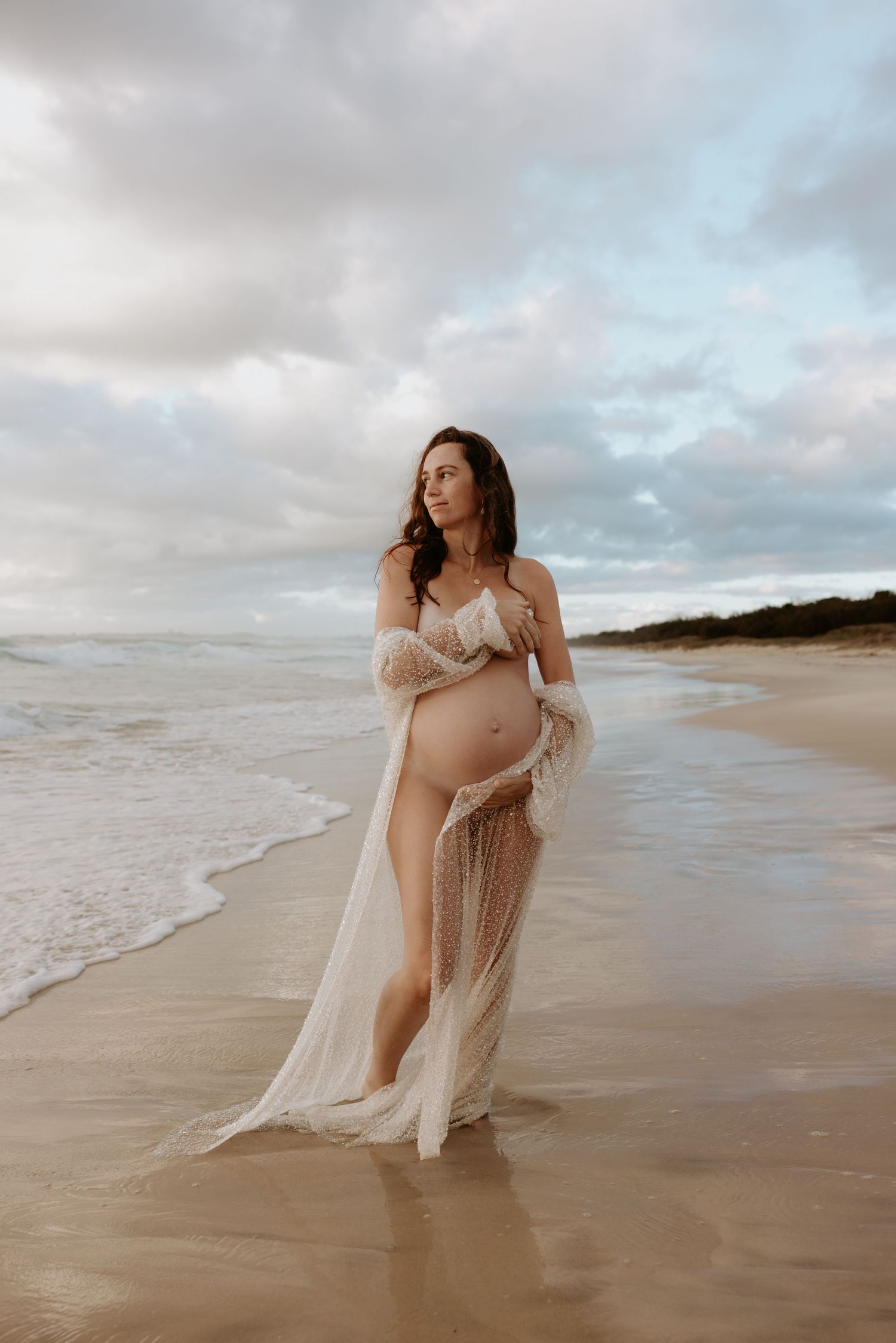 Ethereal maternity photoshoot on a cloudy beach with flowing white sheer dress and gentle waves in the background.