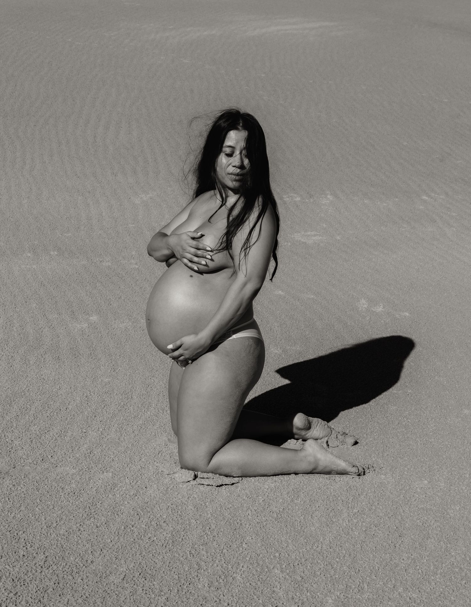 Pregnant mum kneels on sand with her eyes closed and holding her baby in her belly.