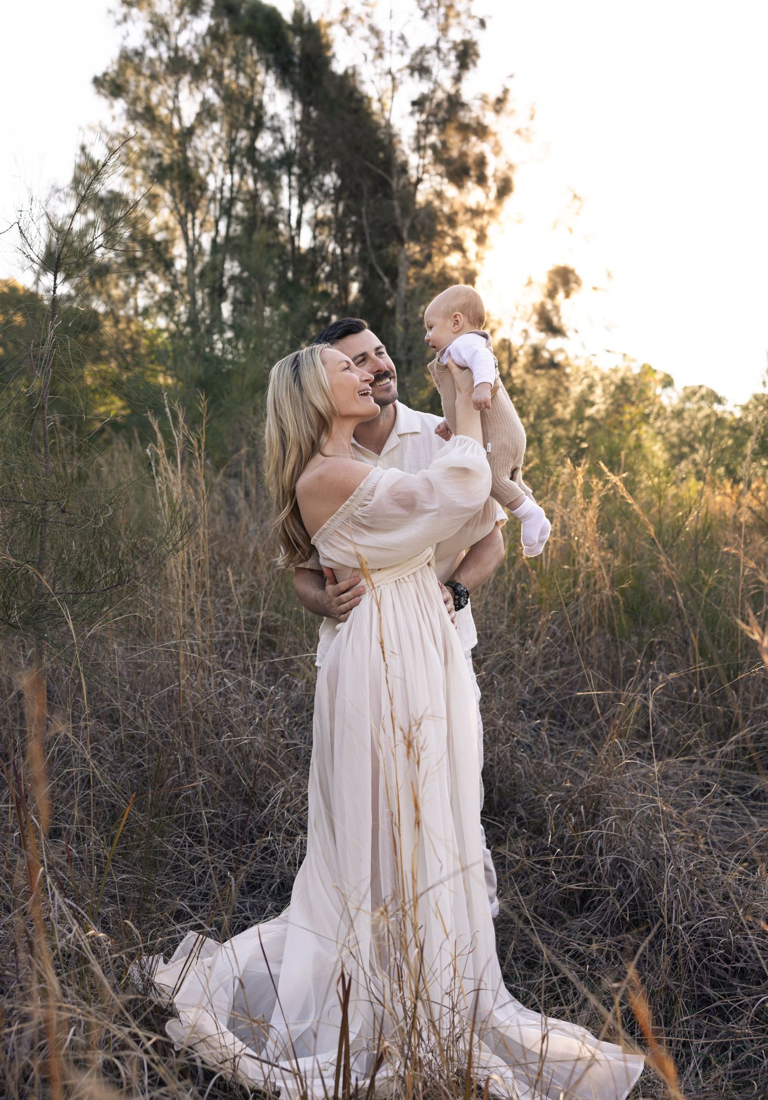 A tender moment captured at sunset with flowing white dresses against tall grass and natural scenery. Mother and father hold their baby high