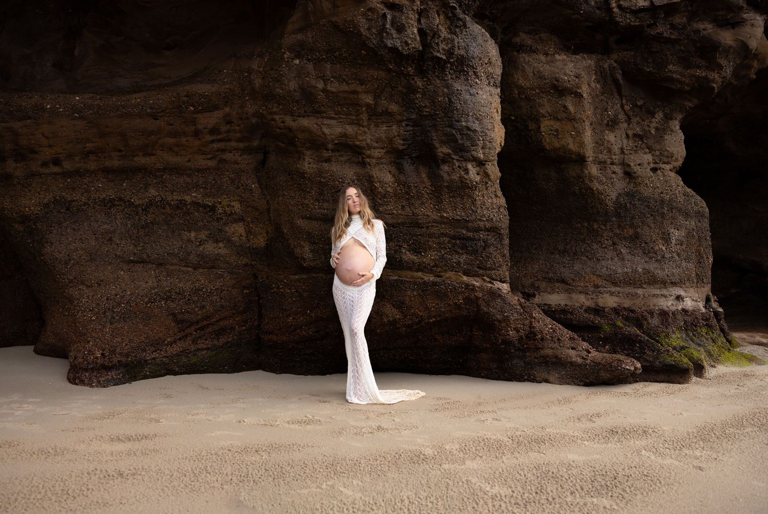 Maternity mother in white dress stands cradling her baby bump, eyes closed looking peaceful with the rocks of Caves Beach behind her.
