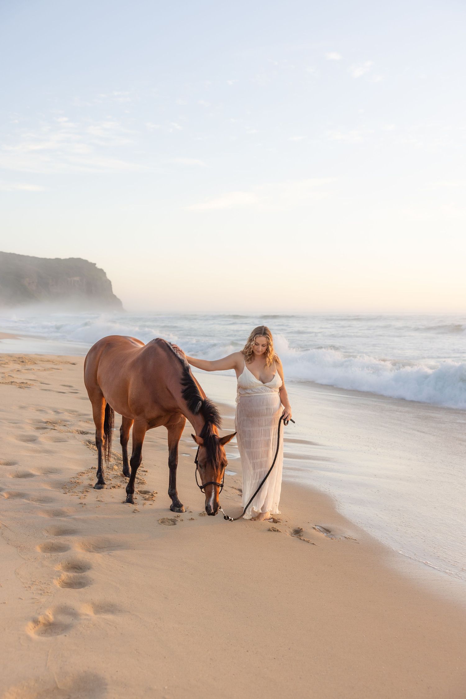 Woman in long white dress leading her horse along the sandy shoreline with waves rolling in behind them and sea mist above the ocean.