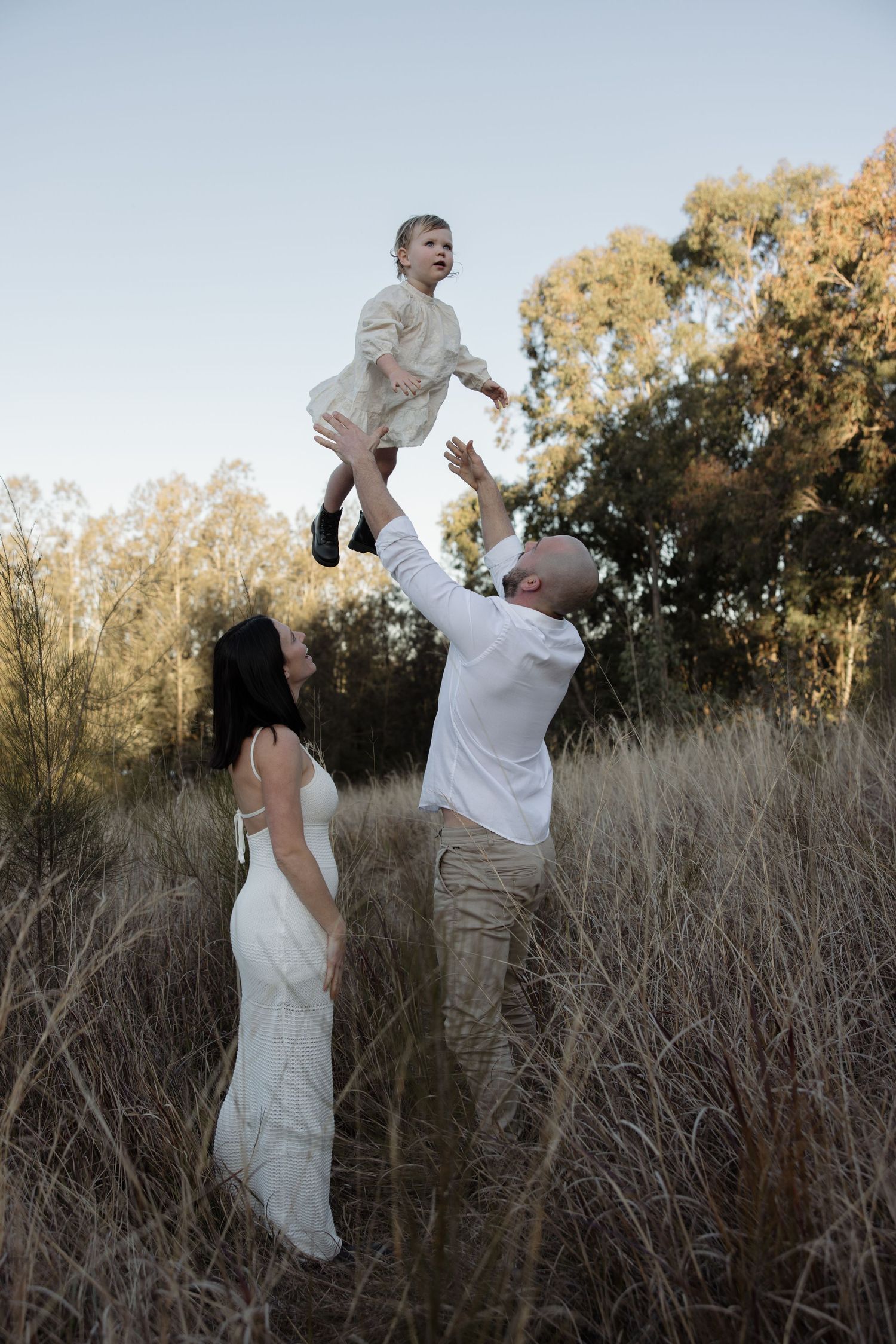 Father plays with daughter throwing her into the air as mother smiles and looks up, holding belly for their second pregnancy announcement.