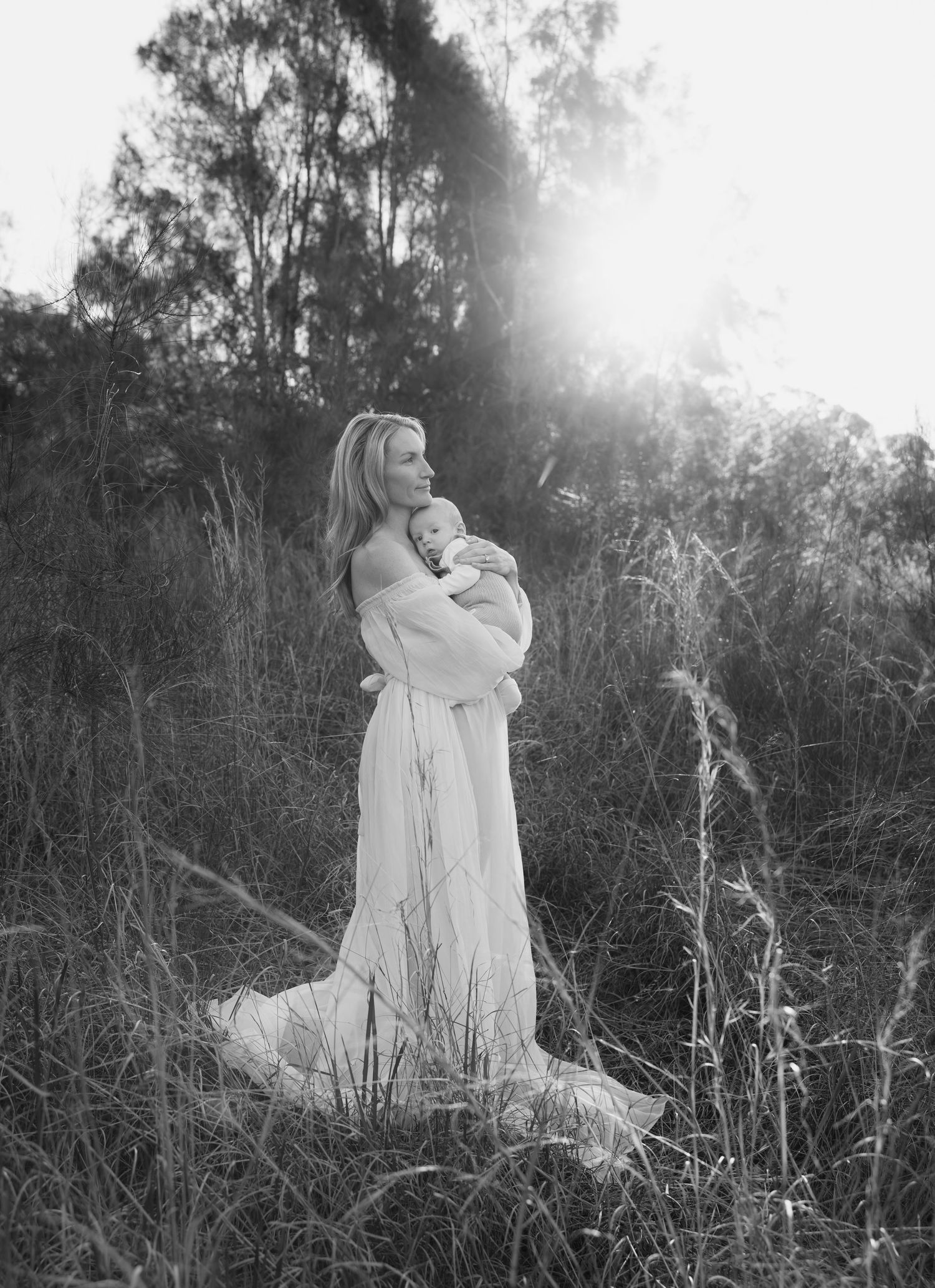 Newborn photography taking place in the tall grass fields with the sun setting behind trees and a mother holding her baby close.