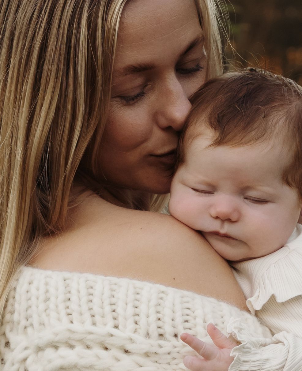 Mother gently kisses sleeping baby on her head. Warms tones are created between their faces and the mother's knit robe.