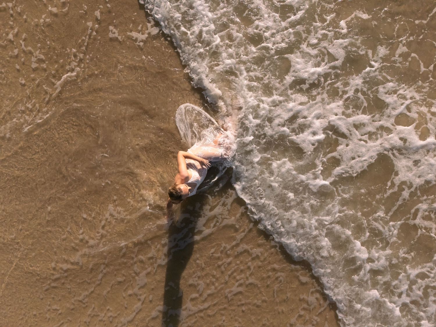 Woman lays in the sand as the drone captures an image from above of a wave rolling in to greet her.