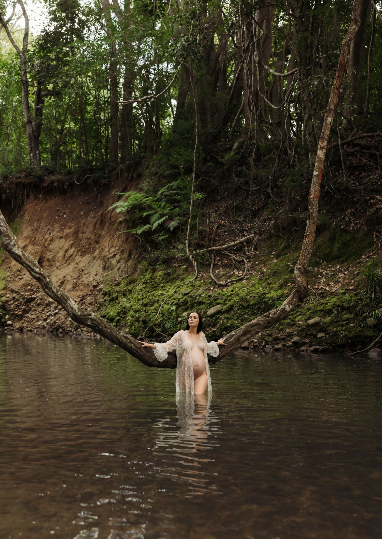 Mother takes an adventure camping in the forest, wading through calm creek waters and looking towards the sun.