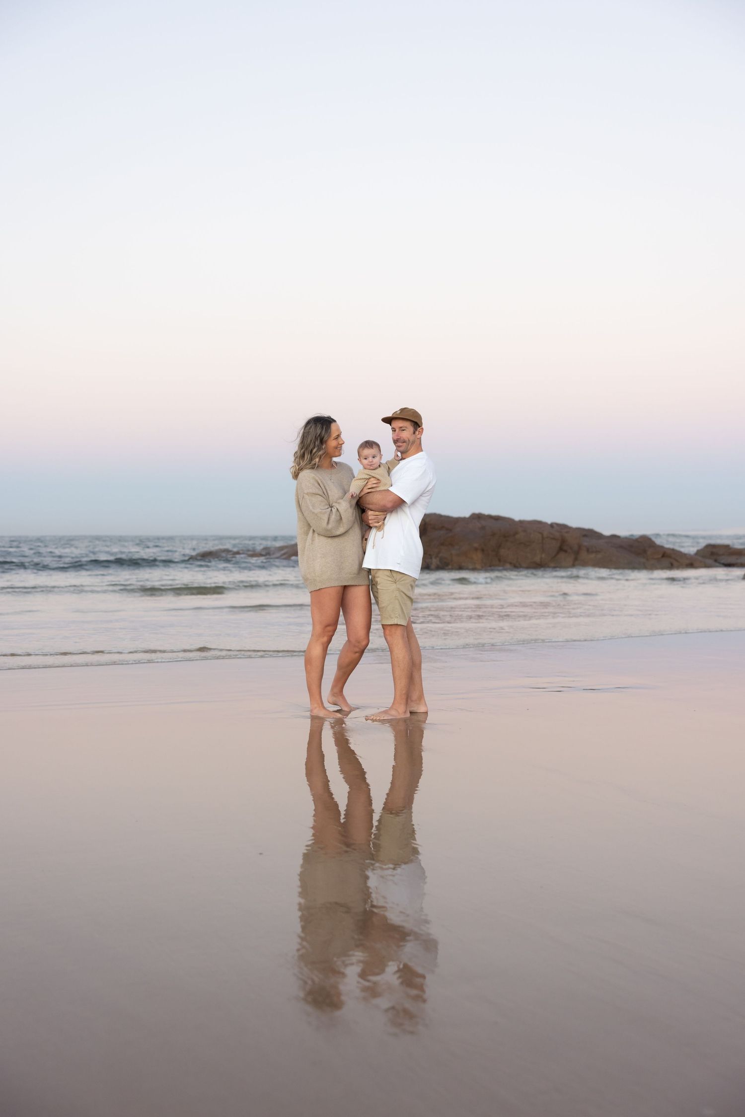 Mum, dad and girl stand at the beach at blue hour in Newcastle. Their reflection is in the smooth water below.
