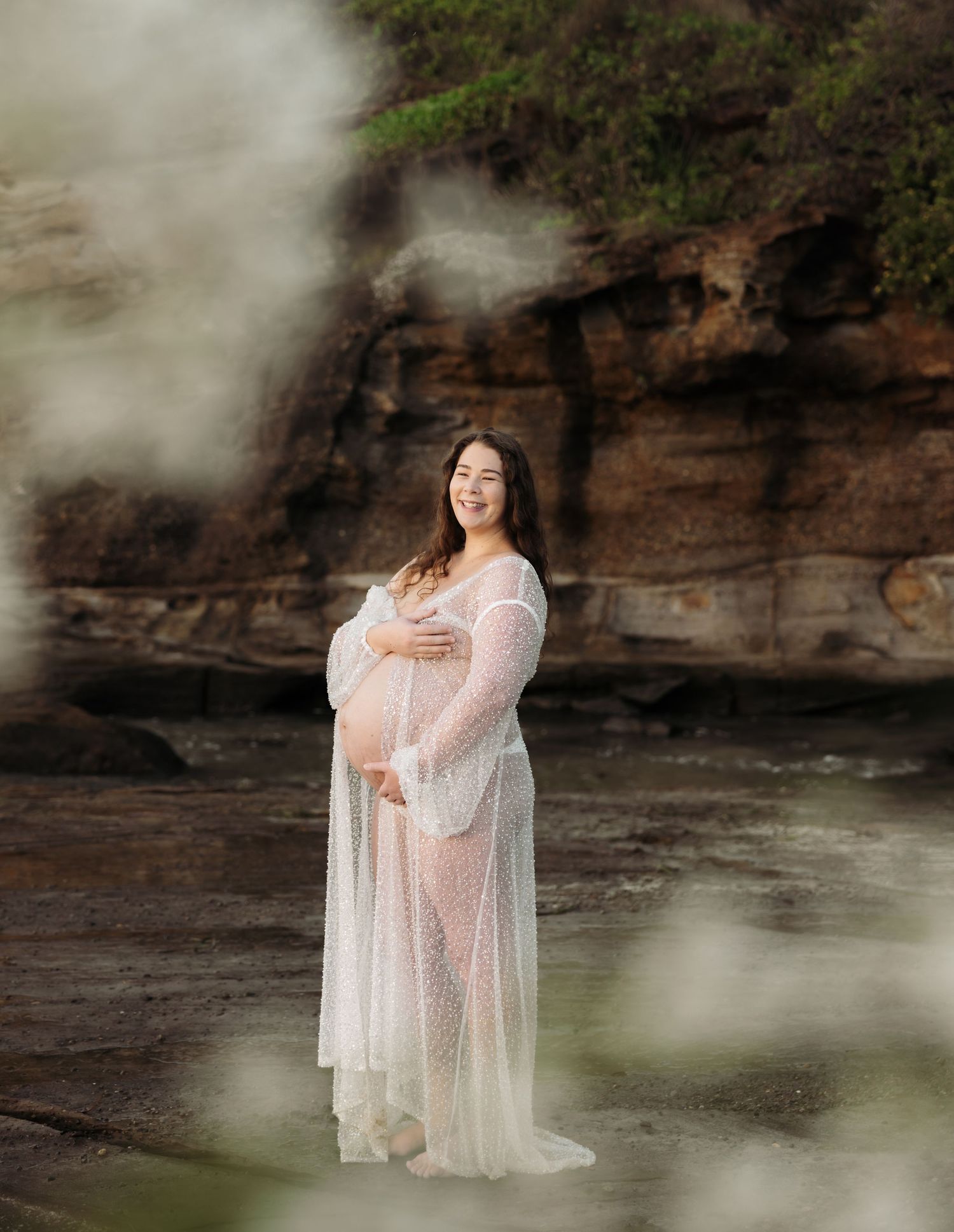 Pregnant mum in sparkle robe is pictured standing infant of rocks. The camera is looking through white flowers creating a hazy effect.
