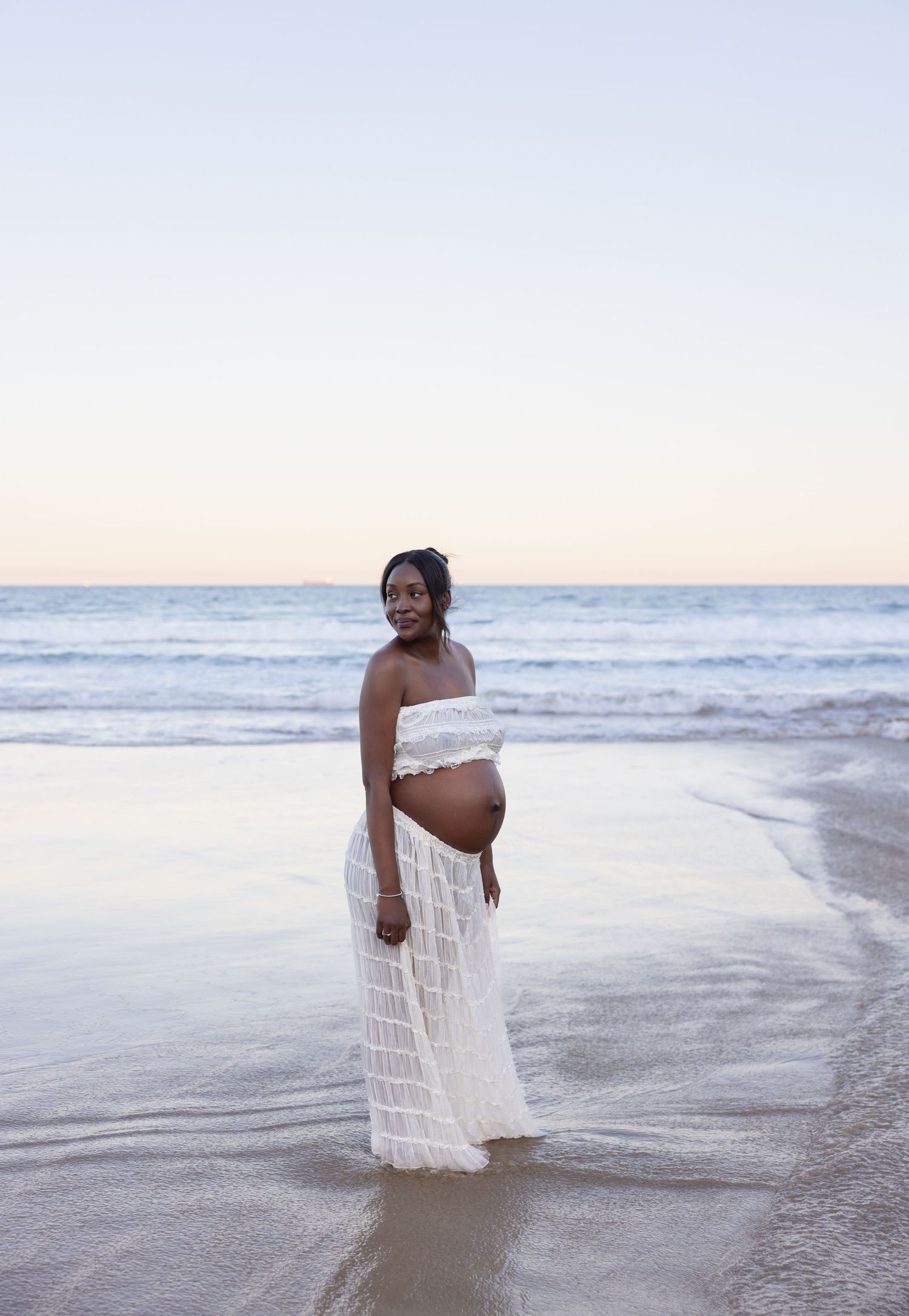Pregnant mum stands still with the blue of the ocean behind her, the clouds reflecting in the shallow water.