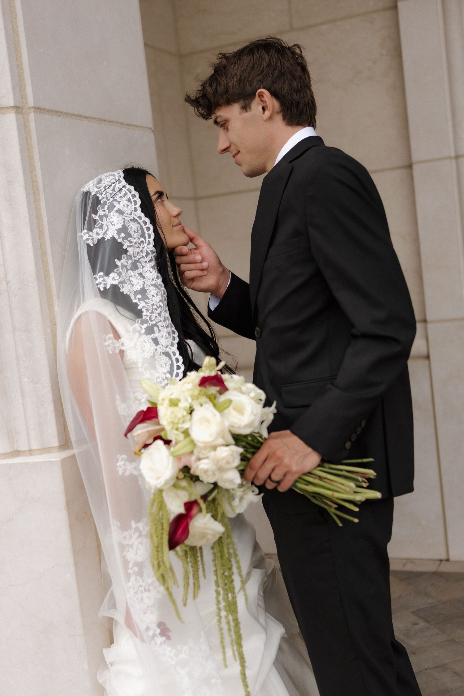 Groom in black tuxedo tenderly touches bride's face as she holds white and red wedding bouquet near stone columns.