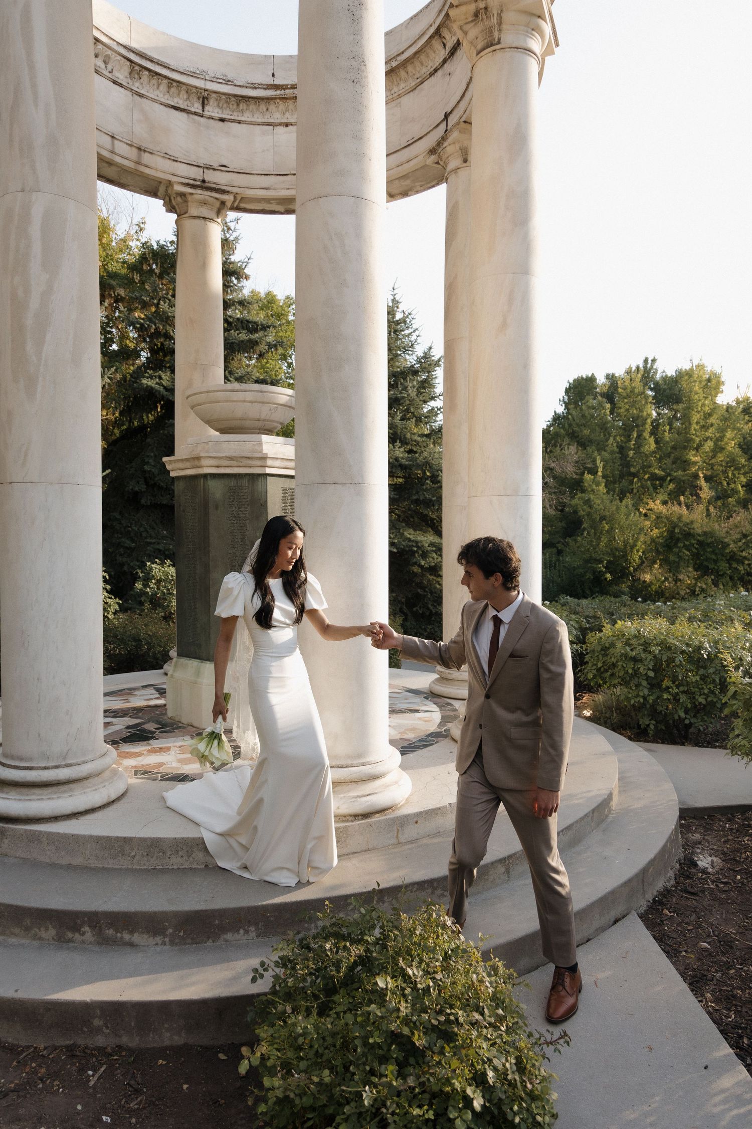 A couple in wedding attire stands beneath a classical white columned gazebo in an elegant outdoor setting.