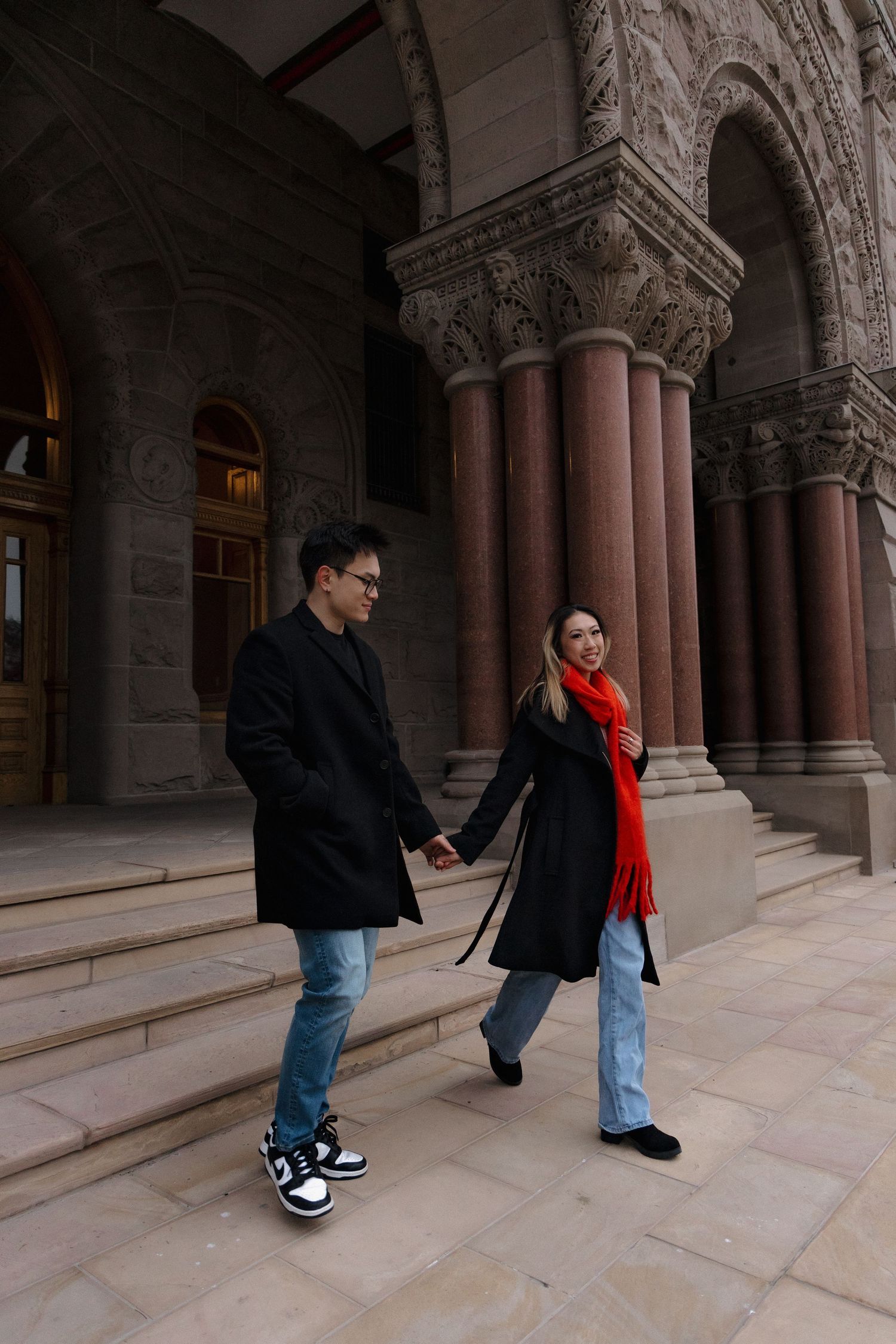 Couple holding hands walking outside ornate stone building with arched columns, woman in red scarf and black coat.