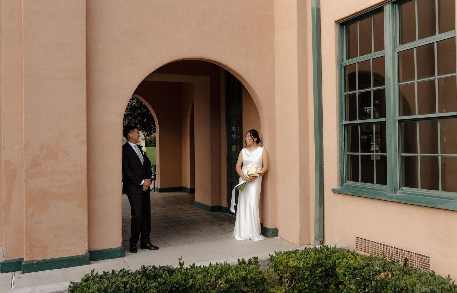 Bride in white gown and groom in tuxedo stand apart under arched colonnade of elegant peach building.