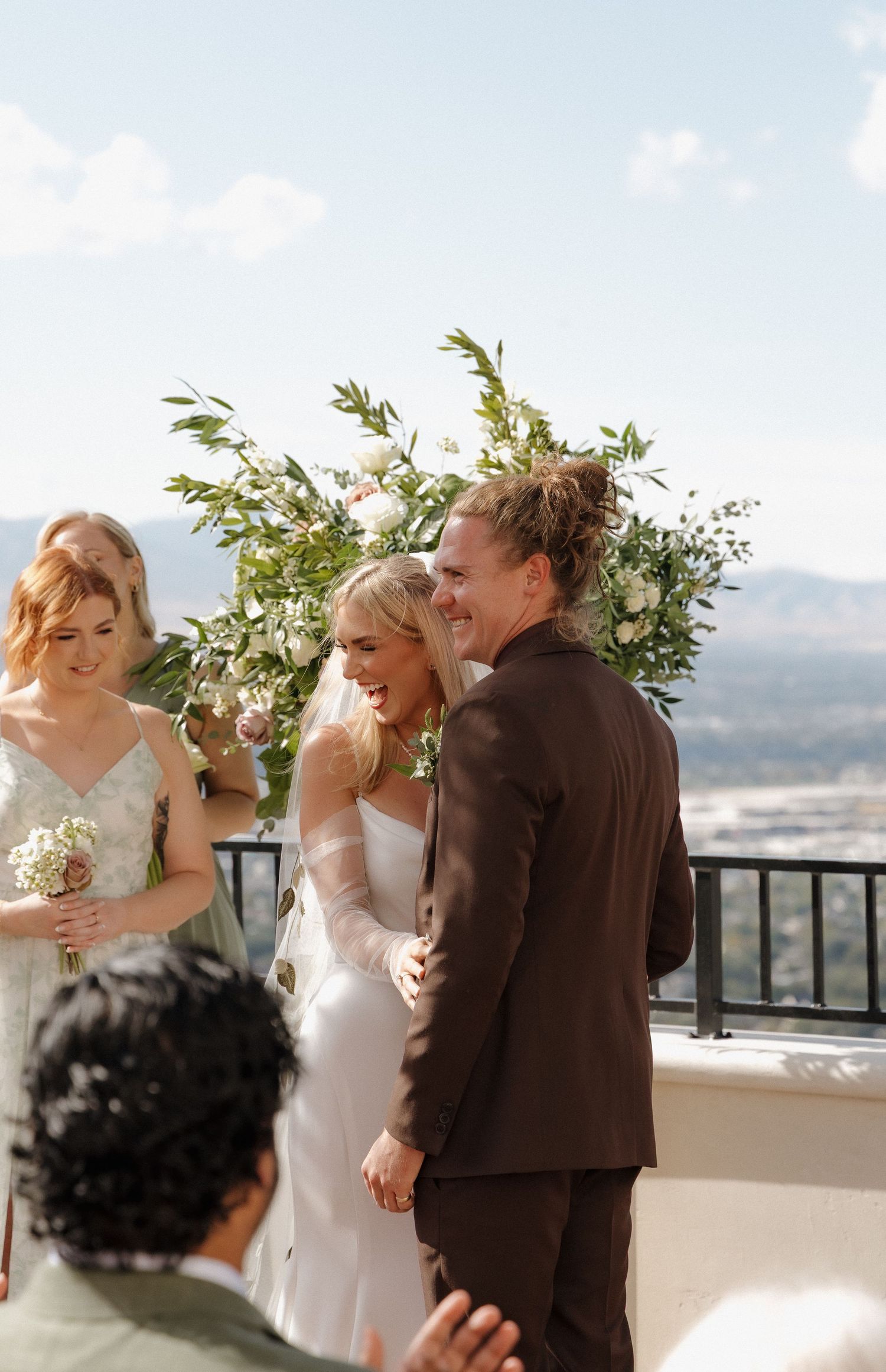 Joyful bride and groom laughing at outdoor wedding ceremony with floral arch and mountain views.
