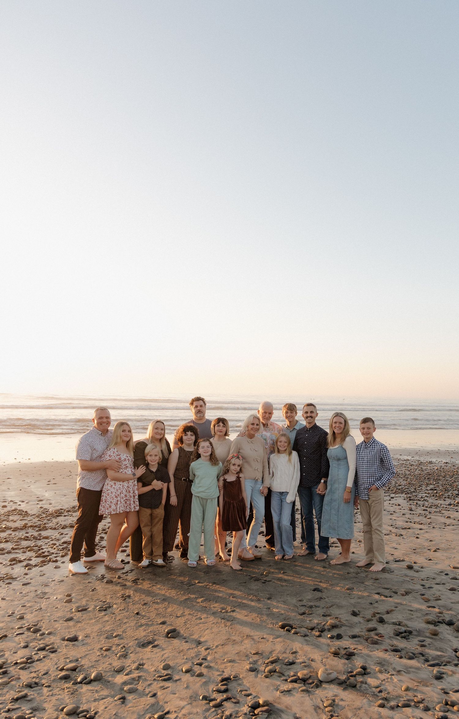 Large family group portrait on a sandy beach at sunset, smiling together by the ocean shore.