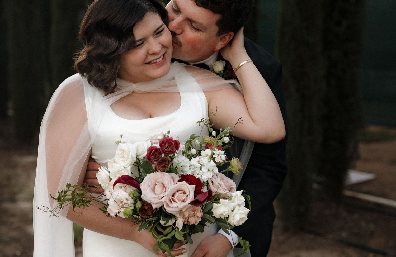 Bride and groom share a tender embrace outdoors, bride holding a bouquet of pink, red, and white roses.