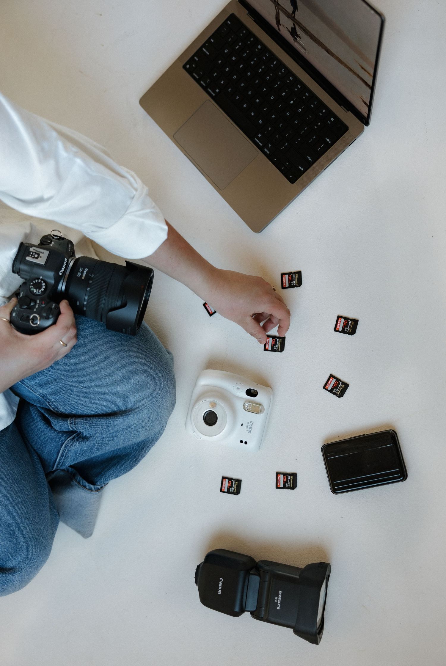Photographer sitting on floor holding DSLR camera surrounded by SD cards, Fujifilm Instax, flash, and MacBook laptop.