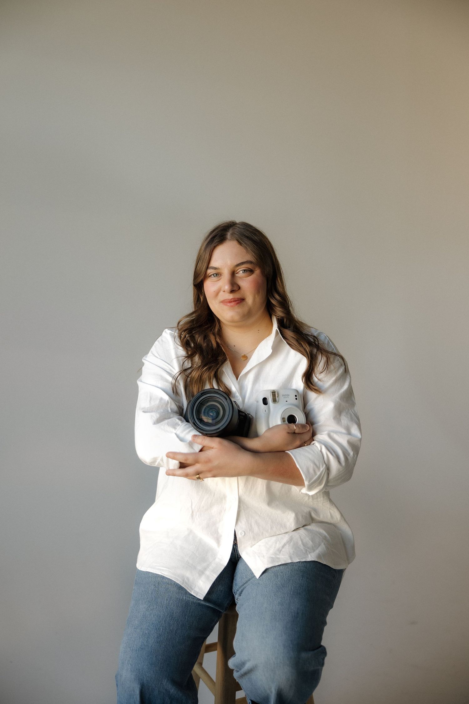 Woman in white shirt and jeans sitting on stool holding a DSLR camera and Fujifilm Instax mini camera.