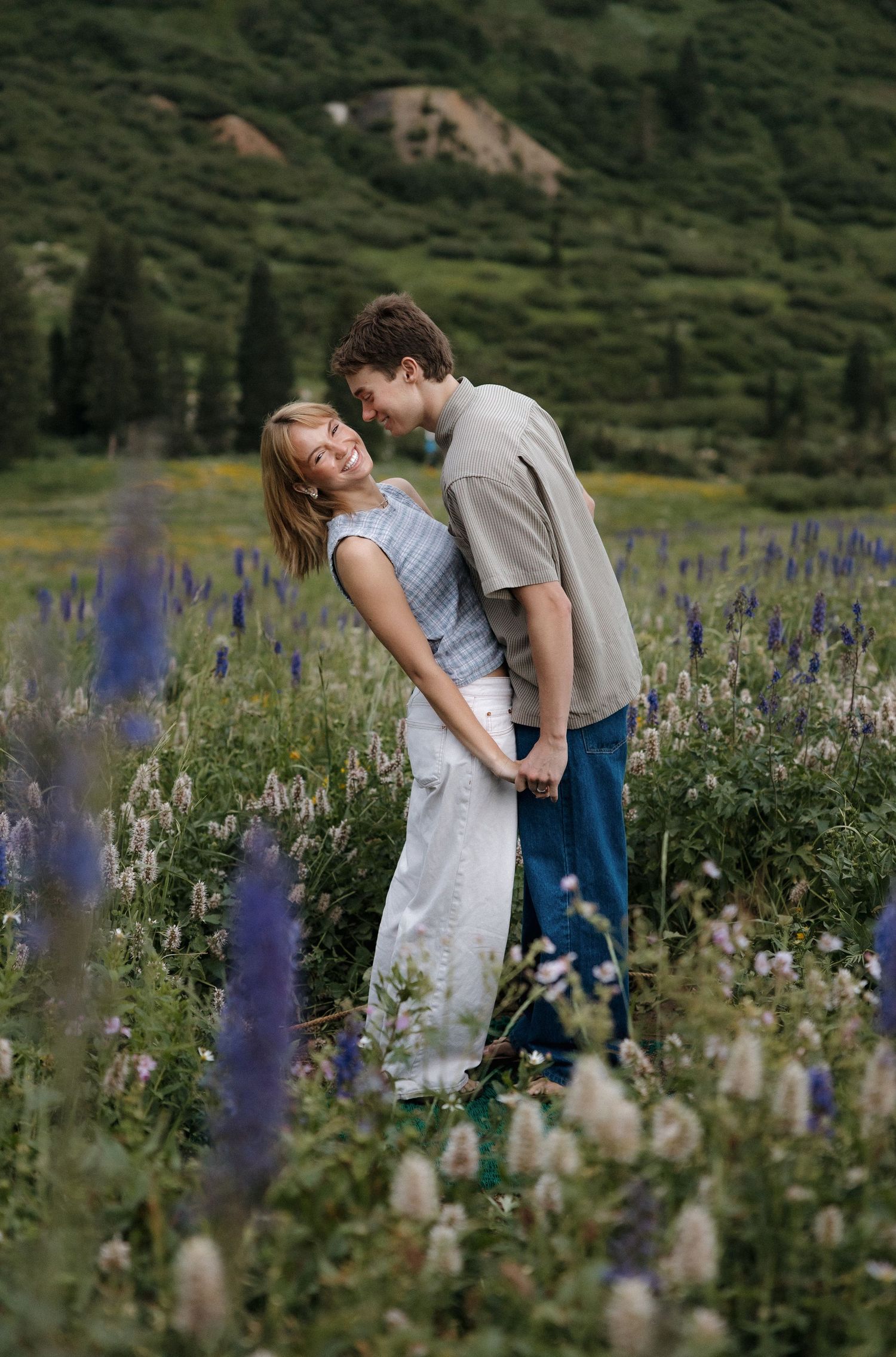 A couple embraces in a lush wildflower meadow with purple lupines, smiling together against a green mountain backdrop.